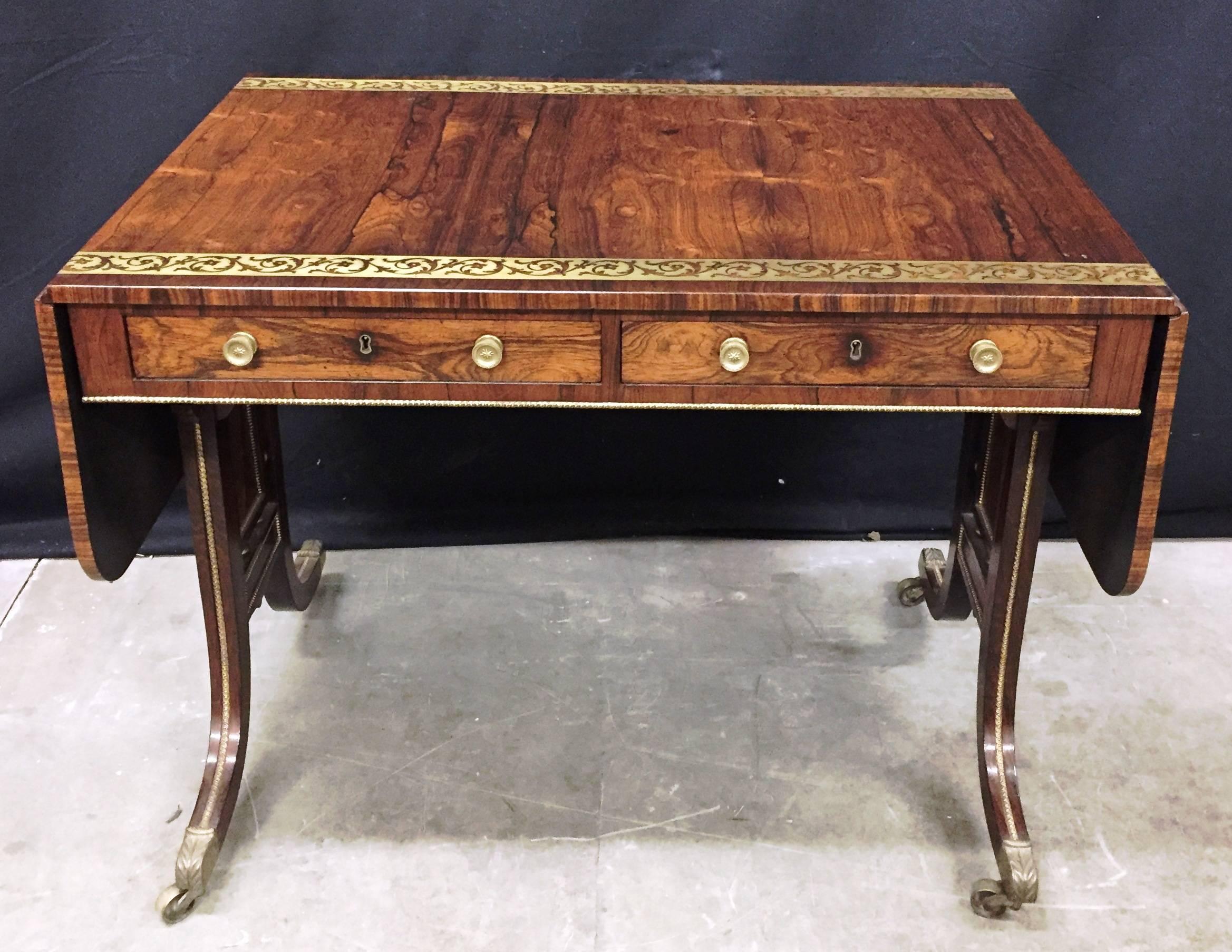 A good quality Regency period Rosewood veneered, brass inlaid sofa table, having two frieze drawers, out swept end supports each with brass mounts and the original brass castors.