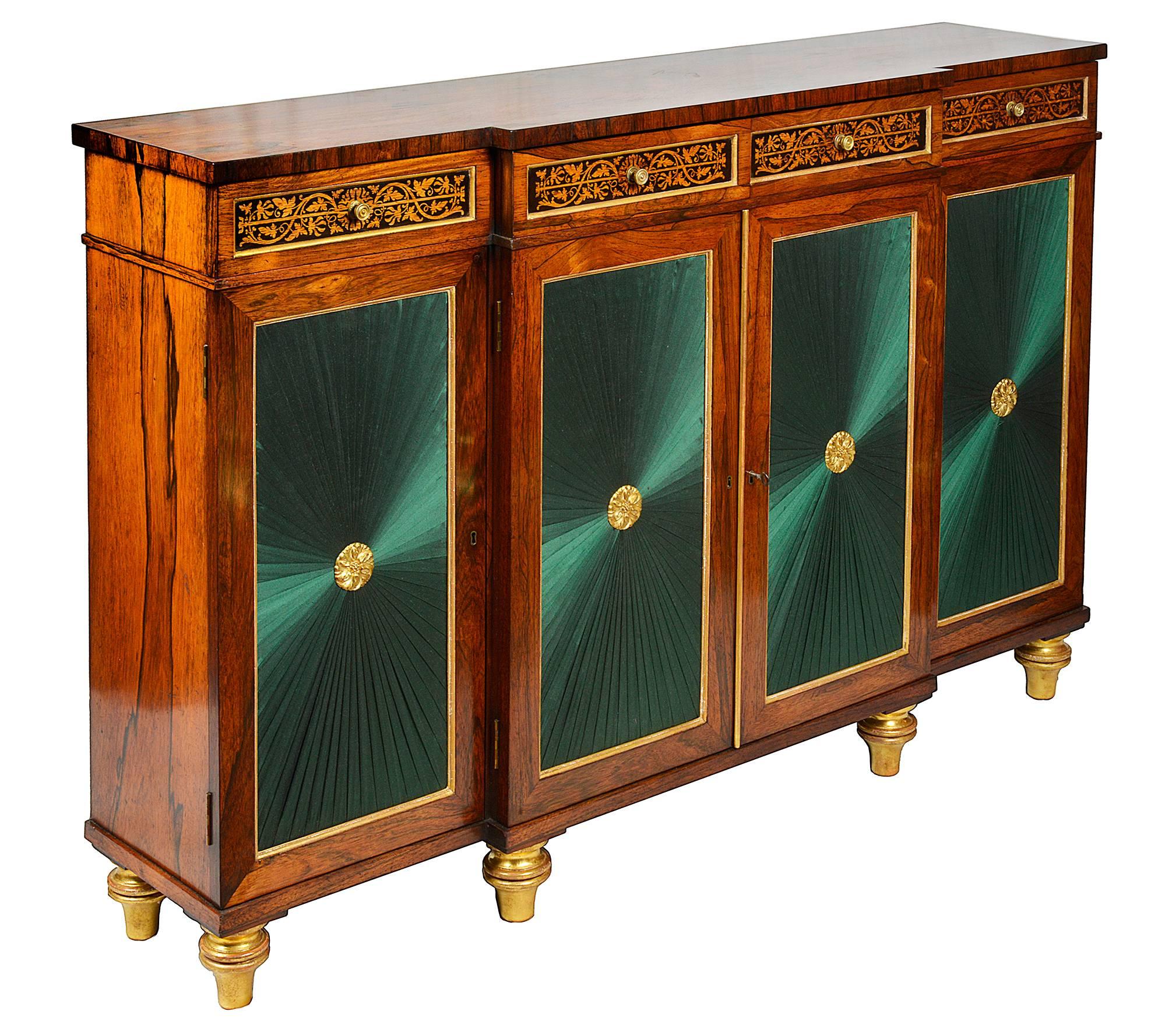 A very good quality Regency period Rosewood break fronted chiffonier side cabinet. Having marquetry inlaid decoration to the drawer fronts, gilded molding and pleated silk panels to the door fronts, raised on turned tapering gilded feet.