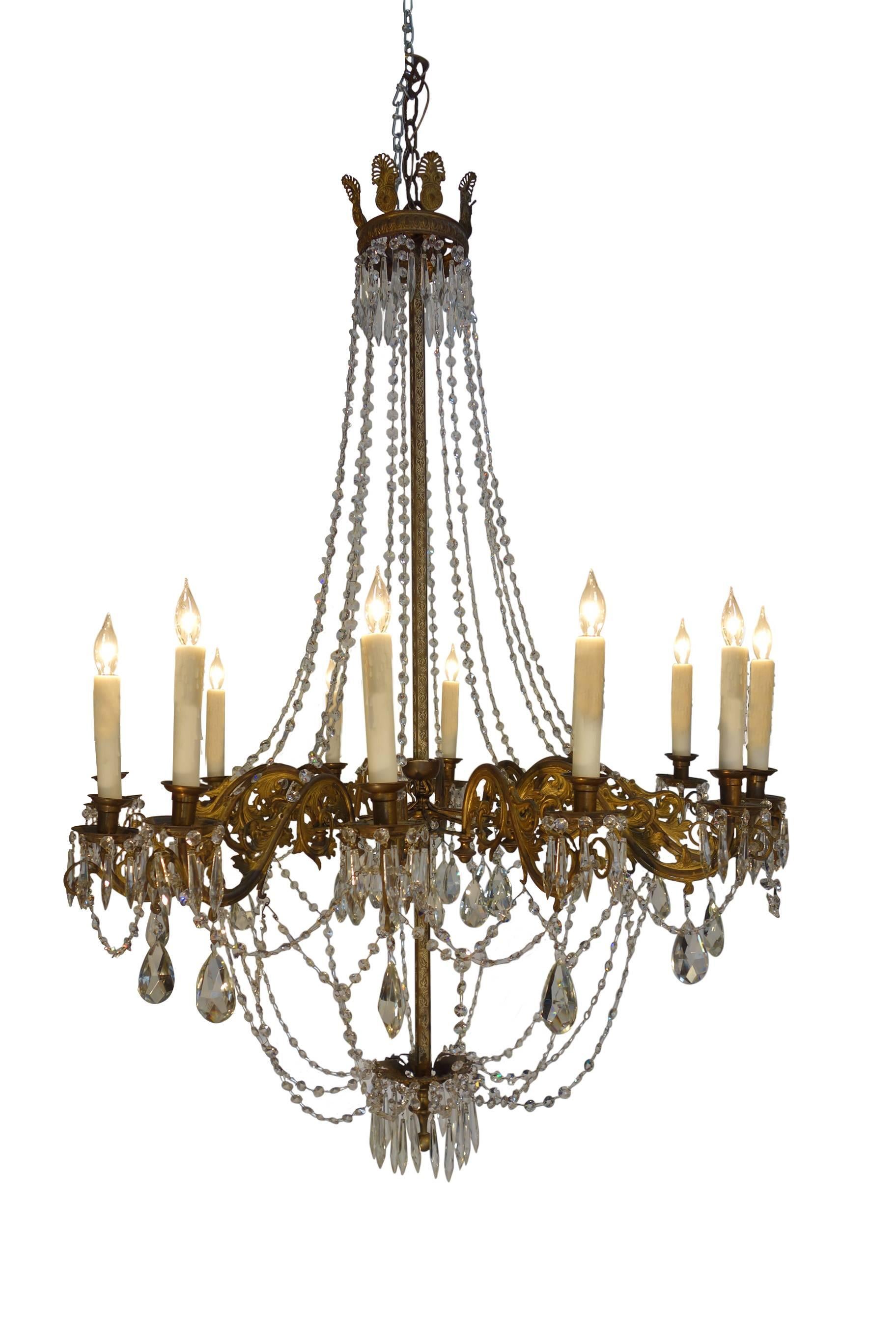 Elegantly tall chandelier, opulent yet stately, featuring 12 lights on brass rocaille arms with crystal pendants, tears and strands. Lovely Empire style crown detail of palms and ivy engraving on the central metal fixture post. Professionally