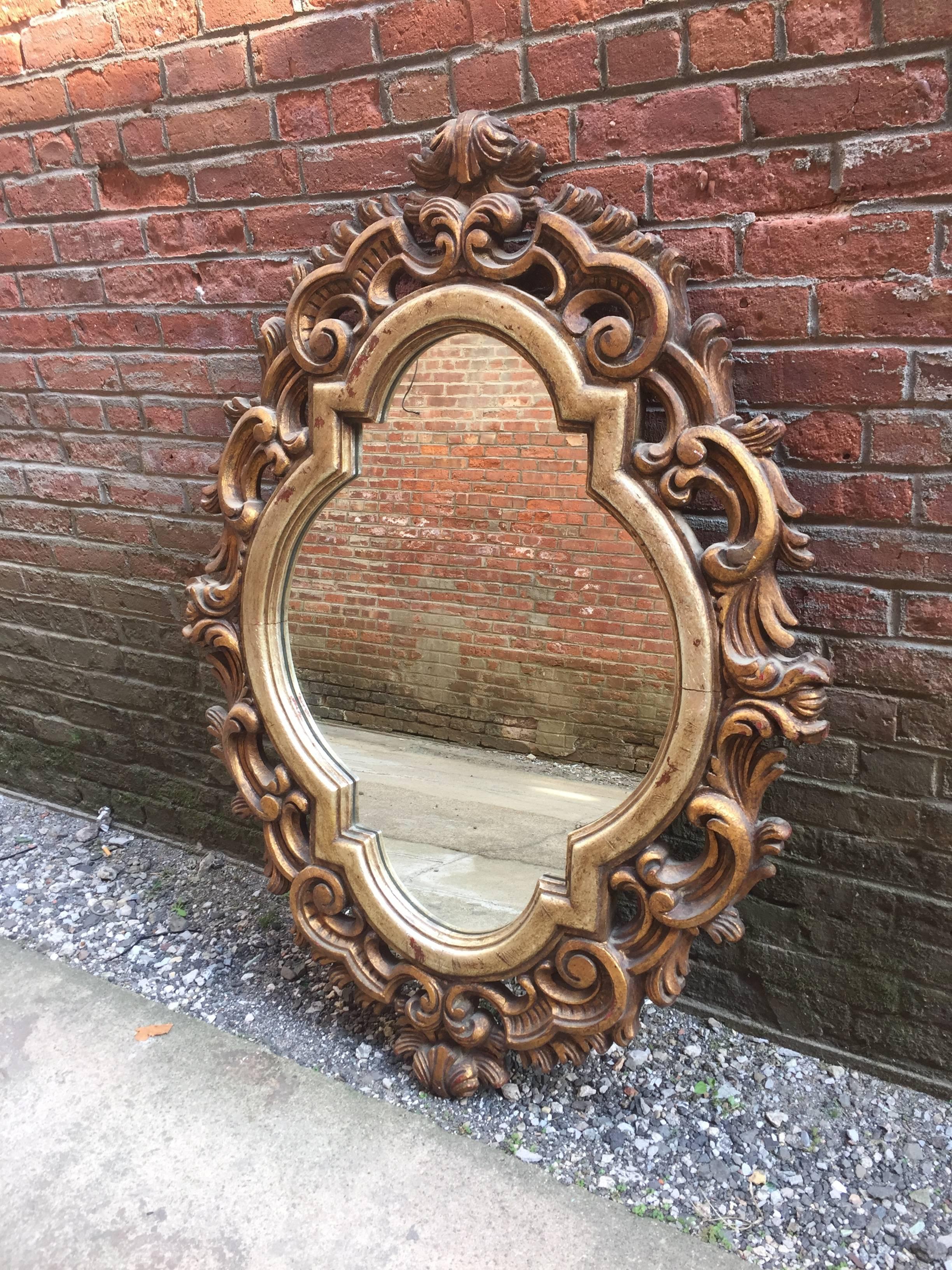 Gilded in gold and silver leaf with red bole peaking through. Heavily carved wood frame surrounding a quatrefoil shaped mirror. Probably Italian origin, circa 1950-1960. The outer wood carved frame is gold leaf gilded and the inner trim is silver