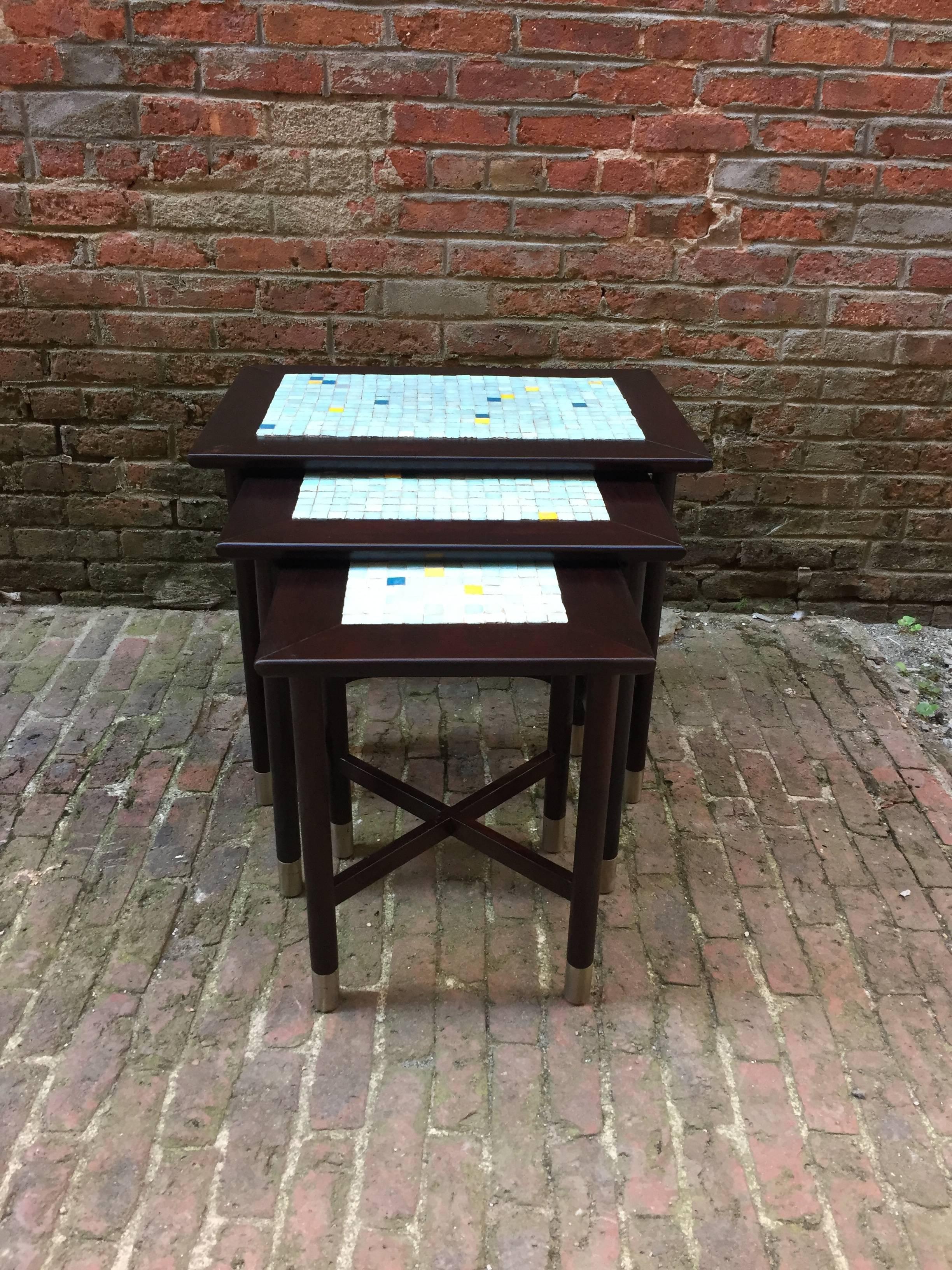 Beautiful set of three wood and glass tile nesting tables in the style of Ed Wormley for Dunbar. Each table has inlaid glass tiles. The colors range from a sky blue, yellow and dark blue. Each mosaic is unique. The round dowel legs end with brushed
