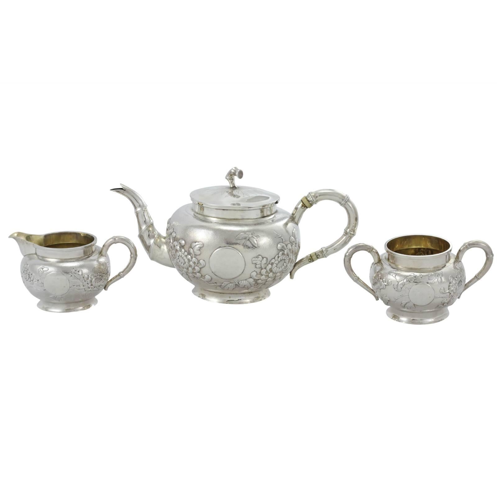 An early 20th century three-piece Chinese silver tea set, decorated with bamboo style handles, chrysanthemums, butterflies and pearls over a hand-hammered surface. Each individual piece has been marked '85', and 'LS', in reference to the purity, and