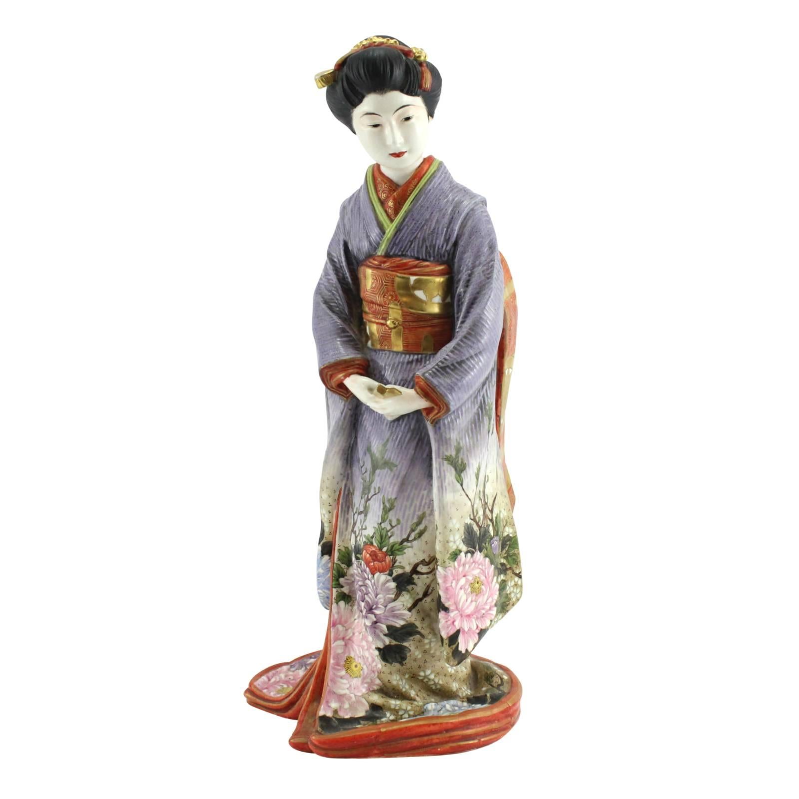 A Japanese Meiji period Awata / Kyoto ware geisha by Kinkozan Sobei. The textured purple kimono of the figure has been hand-painted with pink, blue, purple, and red flowers that flow up the robe. The folds, trim, cuffs, and neck of the kimono are