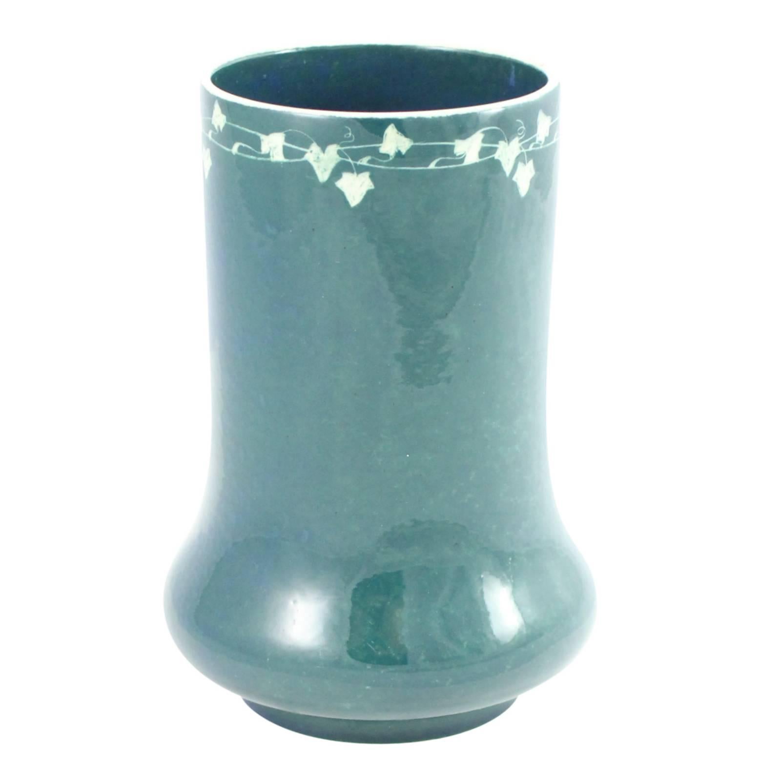 An early 20th century Arts and Crafts ceramic vase by Ruskin Pottery. The piece features a mottled deep teal glaze over a white base, with incised ivy leaves around the rim of the vase. The base of the piece is marked 