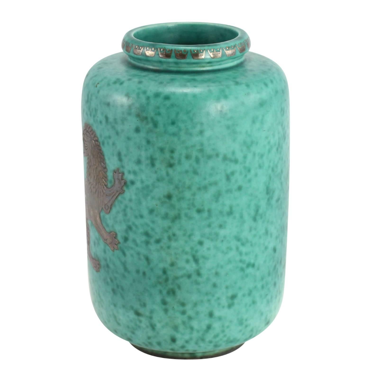 An ‘Argenta’ green stoneware vase with silver overlay by designer Wilhelm Kåge for Gustavsberg, Sweden. Decorated with overlaid silver crowns along the rim and a silver lion in the centre of the piece; this mottled green vase was apart of Kåge’s