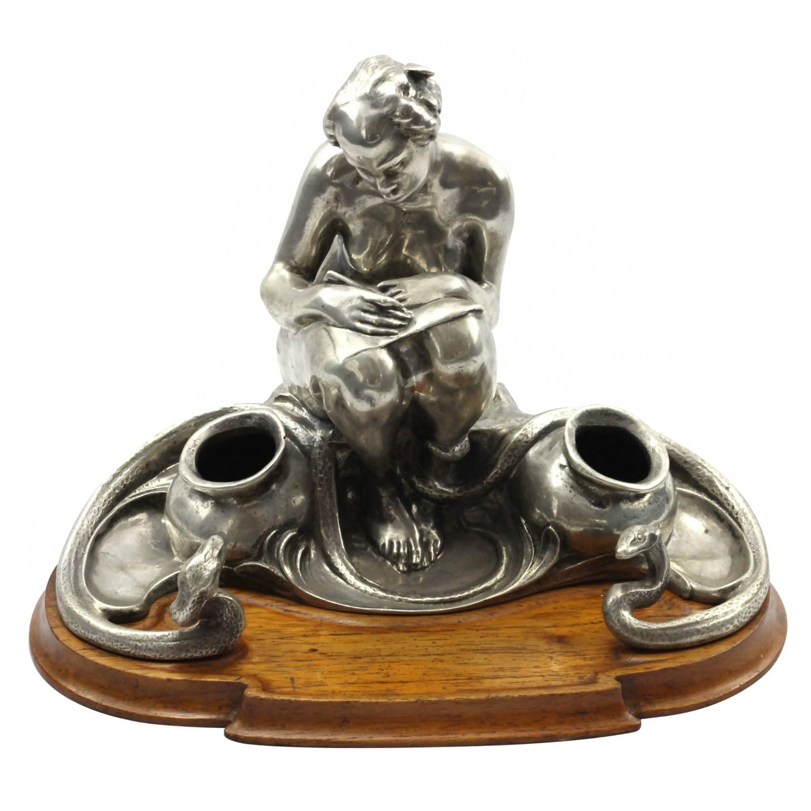 An early 20th century Art Nouveau .800 continental silver inkstand. The piece depicts a nude woman resting a piece of paper on her lap, with a pen in her hand. Beside her are two inkwells, and two serpents, one of which has its tail wrapped around