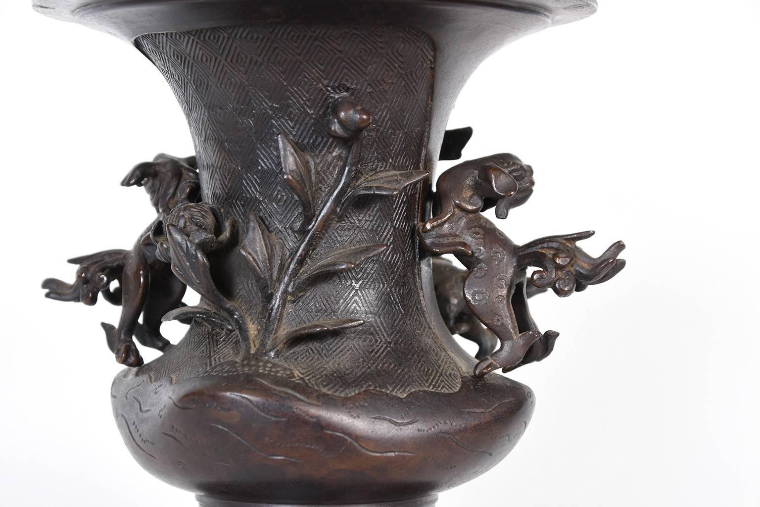 Antique Japanese cast bronze offering vase with removable shi shi handles. There are two additional shi shi dogs playing amongst flora on the front, and a flower stem on the back. The background is filled with an etched geometric diamond