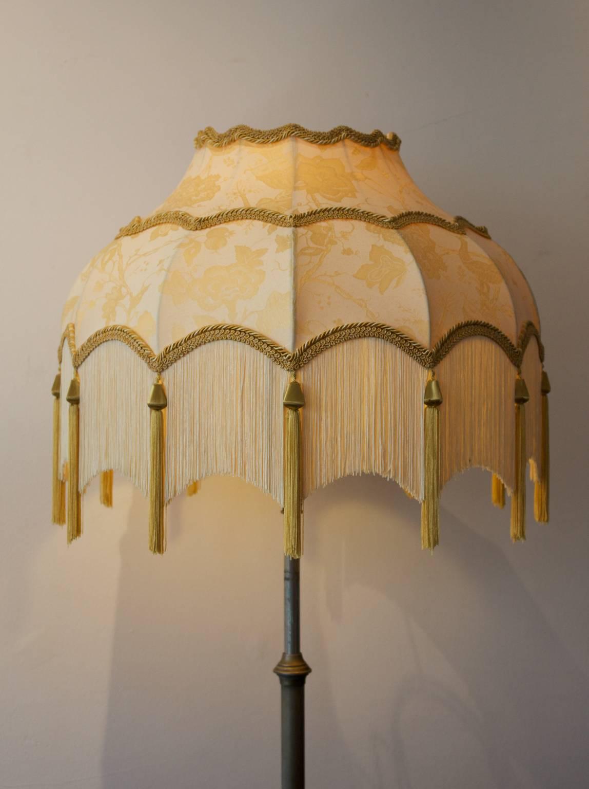 An arts and crafts lamp with large fringed shade. Early 20th century, England.

Figured lamp base of brass plated steel on tripod legs, with flamboyant fabric shade - later work - very nicely made with good lining and gold fringes. 

The lamp base