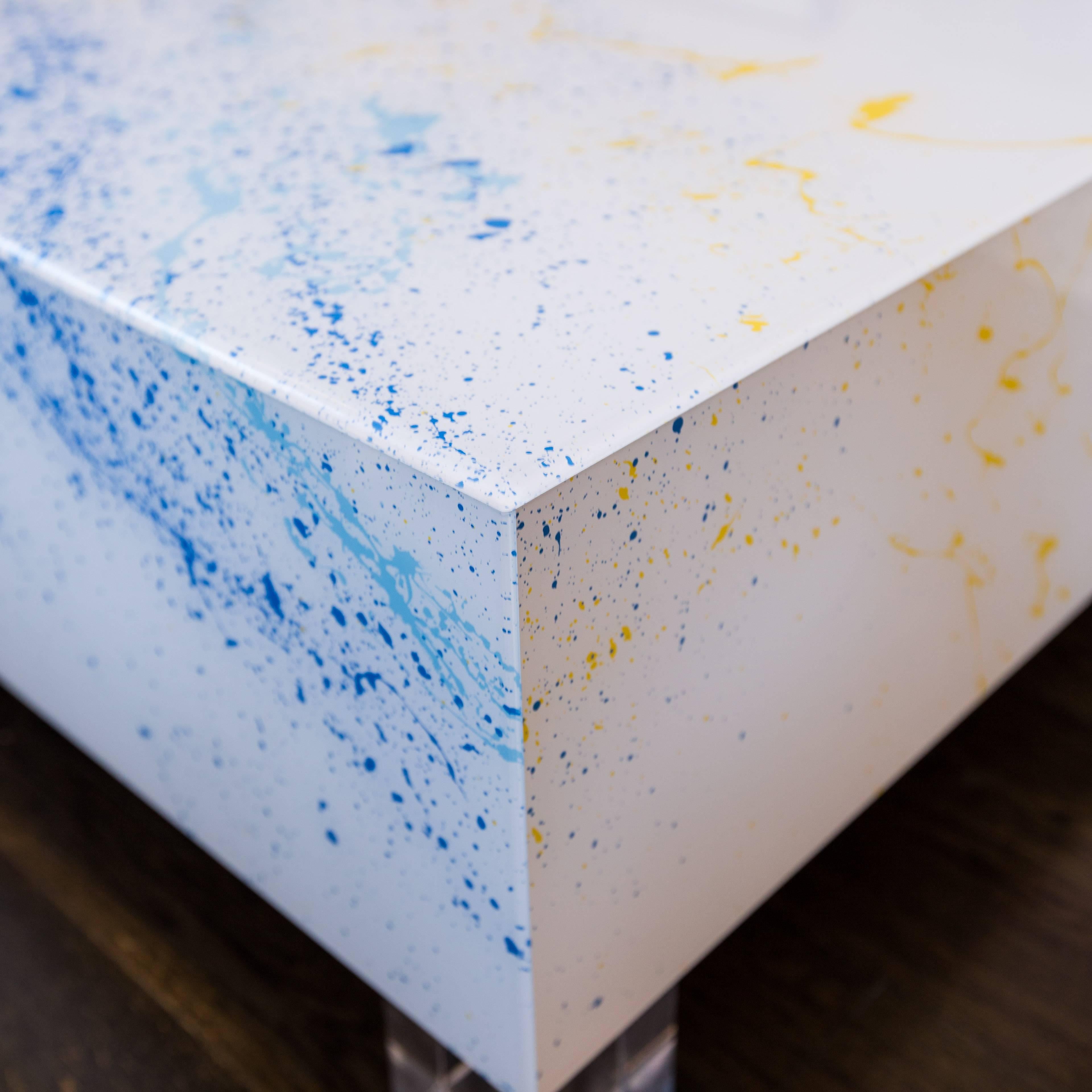 Custom, one-of-a-kind coffee table made of reverse drip-painted Lucite panels sitting on solid Lucite feet. White background with two shades of blue and some yellow Jackson Pollock-like splatters.