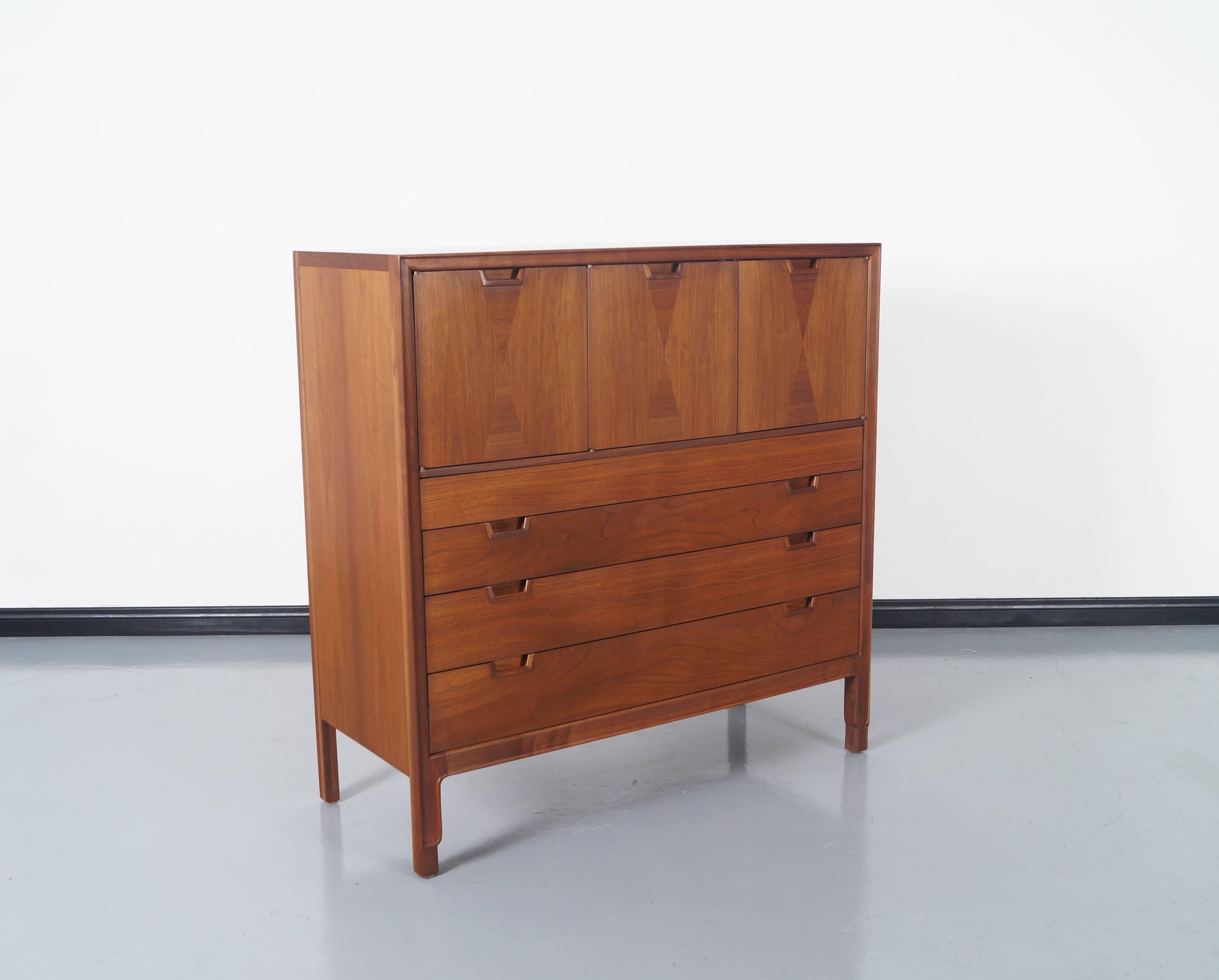 Mid-Century walnut highboy by John Stuart for Janus Collection. Features three compartments on top and four drawers on the bottom. Beautiful inlaid details on the top doors.