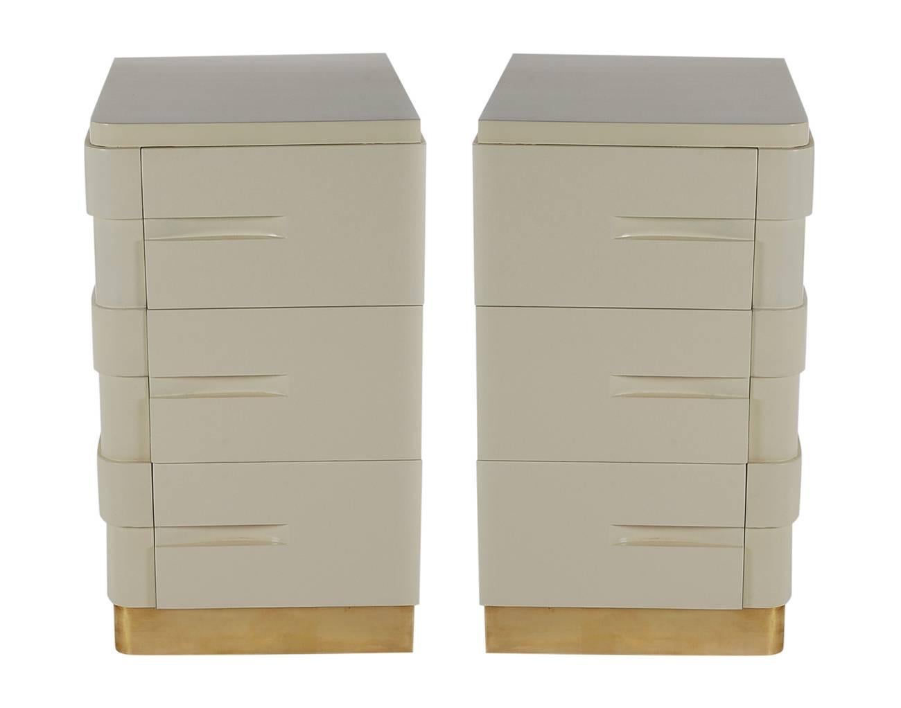 A handsome pair of American Art Deco nightstands, circa 1940s. They feature solid wood construction, newer bone white lacquer, and brass platform bottoms. 

In the style of: Gilbert Rohde, James Mont and Paul Frankl.