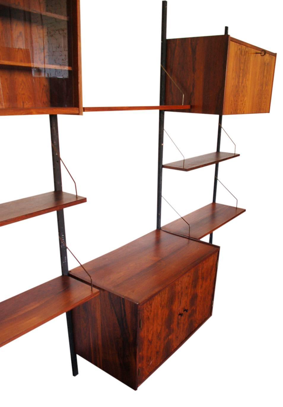 A three bay rosewood PS wall system designed by Peter Sorensen for Randers, circa 1960s. It features one pull down door bar cabinet, one glass display case with two shelves, one two door cabinet with interior shelf, and five floating shelves. All