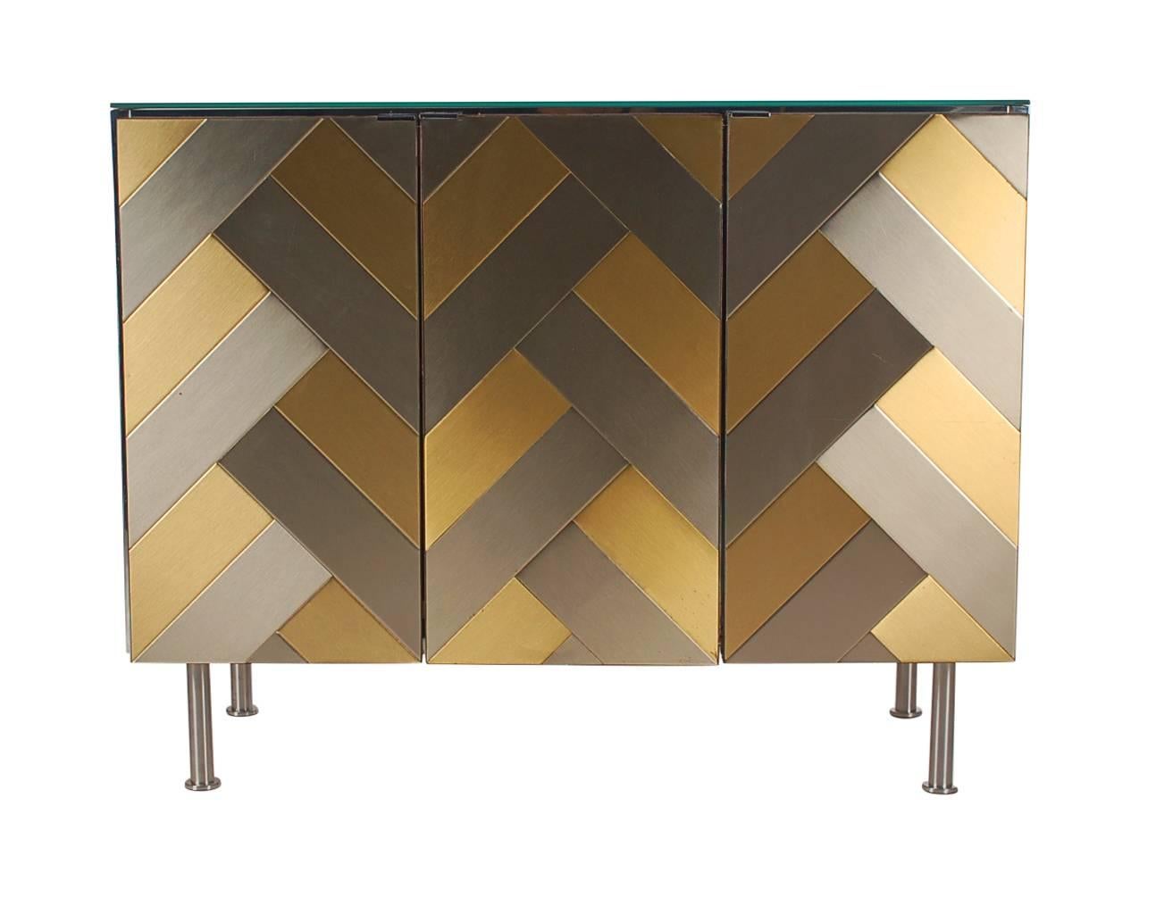 A beautiful interior lit bar cabinet by Ello Furniture Company. It features a brass and steel herringbone front, mirrored top and lighted white laminate interior.