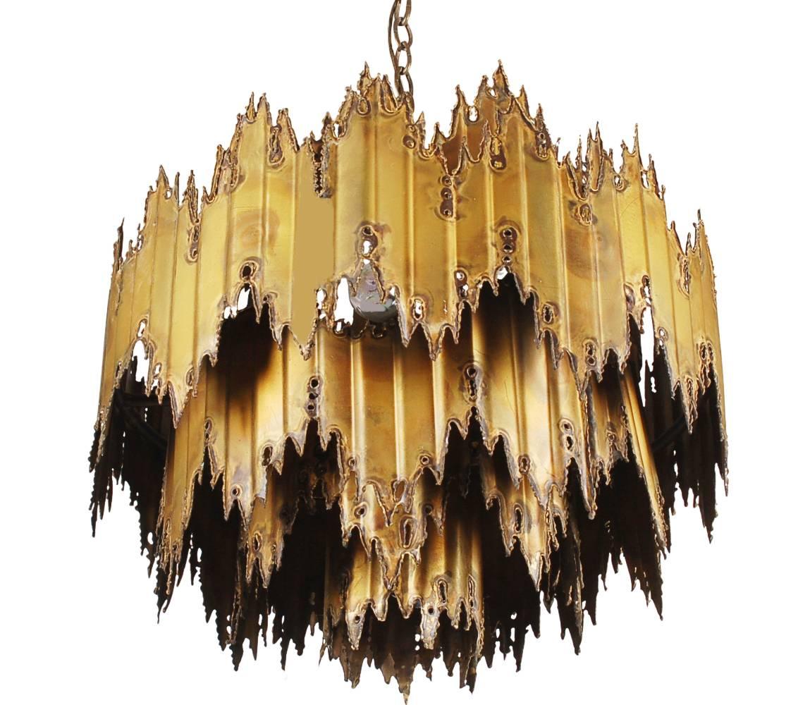 An incredible torch cut brass chandelier designed by Tom Greene and produced by Feldman Lighting. It is fully working with the original ceiling plate. Manufacturers label is present. 

In the style of: Tom Green, Curtis Jere, Silas Seandel, Paul