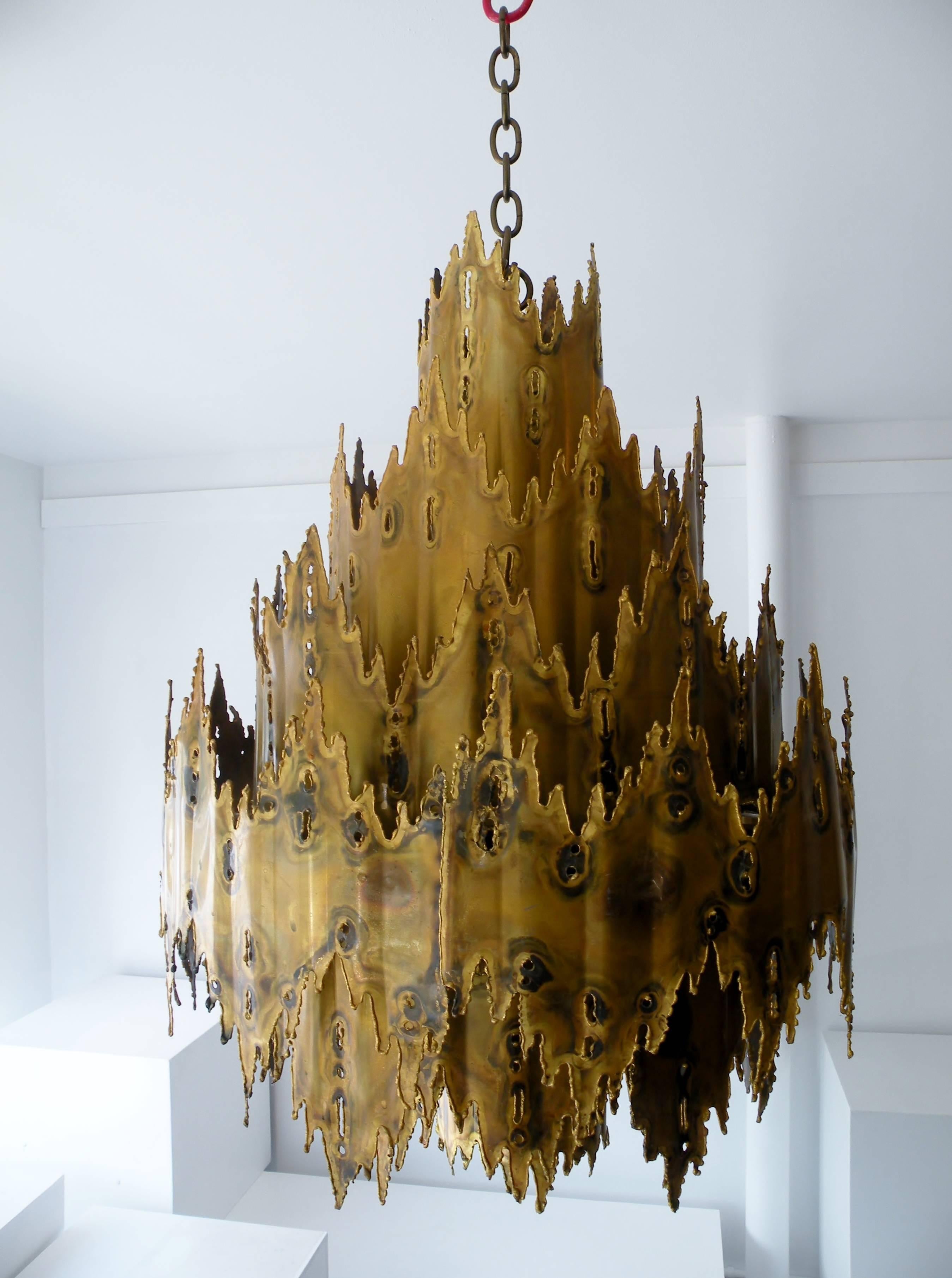 The iconic Brutalist chandelier designed by Tom Green for Feldman Lighting. This is not the wallflower in the room with impressive scale and striking design. Constructed with four tiers of torch cut metal with a warm patinated brass surface.