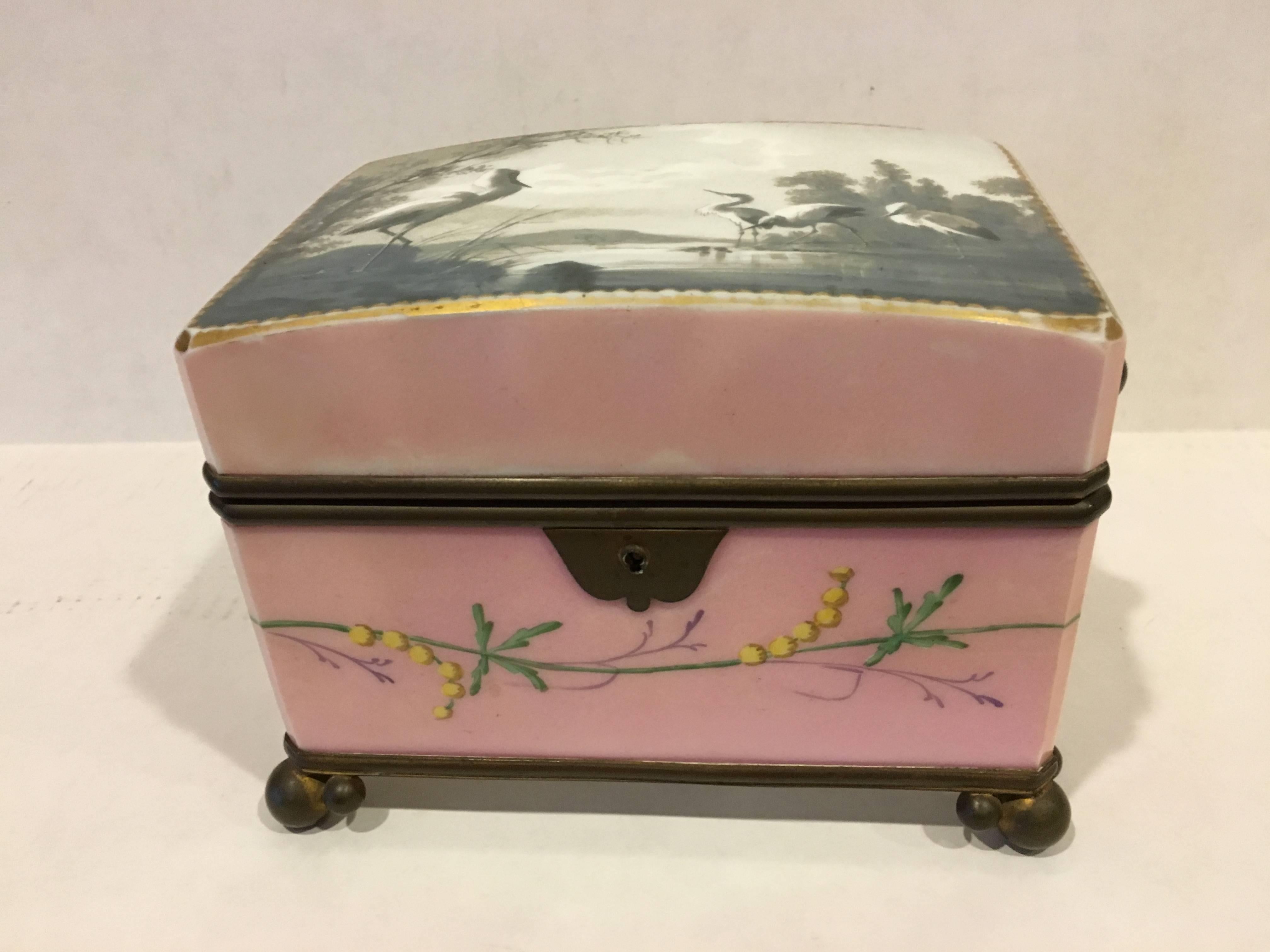 A beautiful hand painted porcelain pink casket box with a floral design all around the box and a grey and white water scene with egrets on the top.
