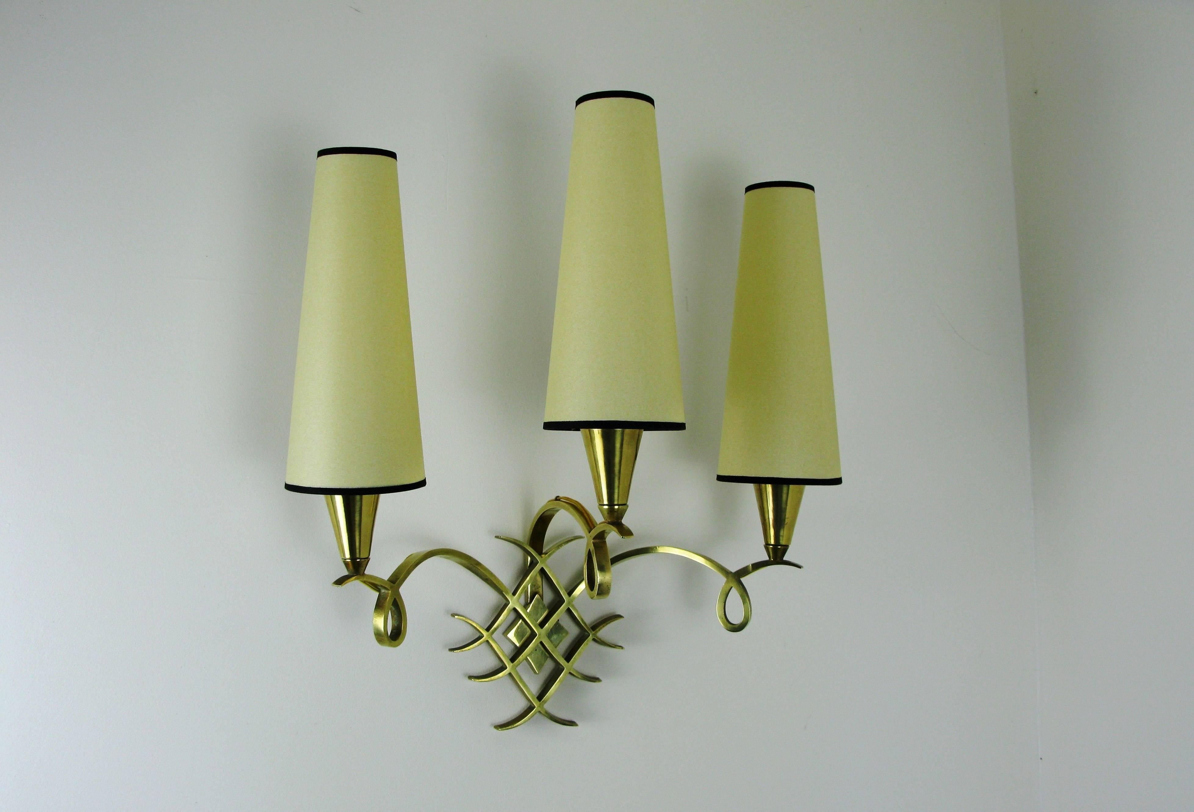 Large pair of polished bronze sconces consisting of a school of three polished bronze lighted arms surmounted by three brass cups holding a conical lampshade bordered with a black line.
The arms are joined to a bronze wreathed mounting.
Lampshades
