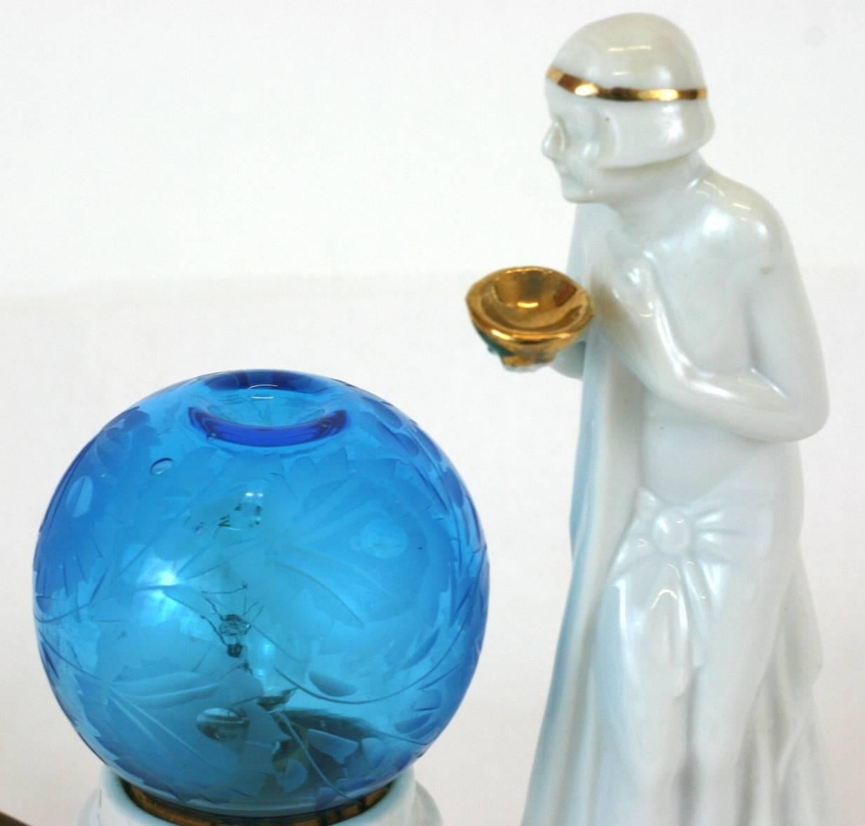 Art Deco porcelain perfume lamp with original blue etched globe. The semi-nude maiden holds a gold cup over an etched blue glass globe, with an indentation to drop perfume oils.
The candle bulb inside the porcelain piece heats the oil to emit the