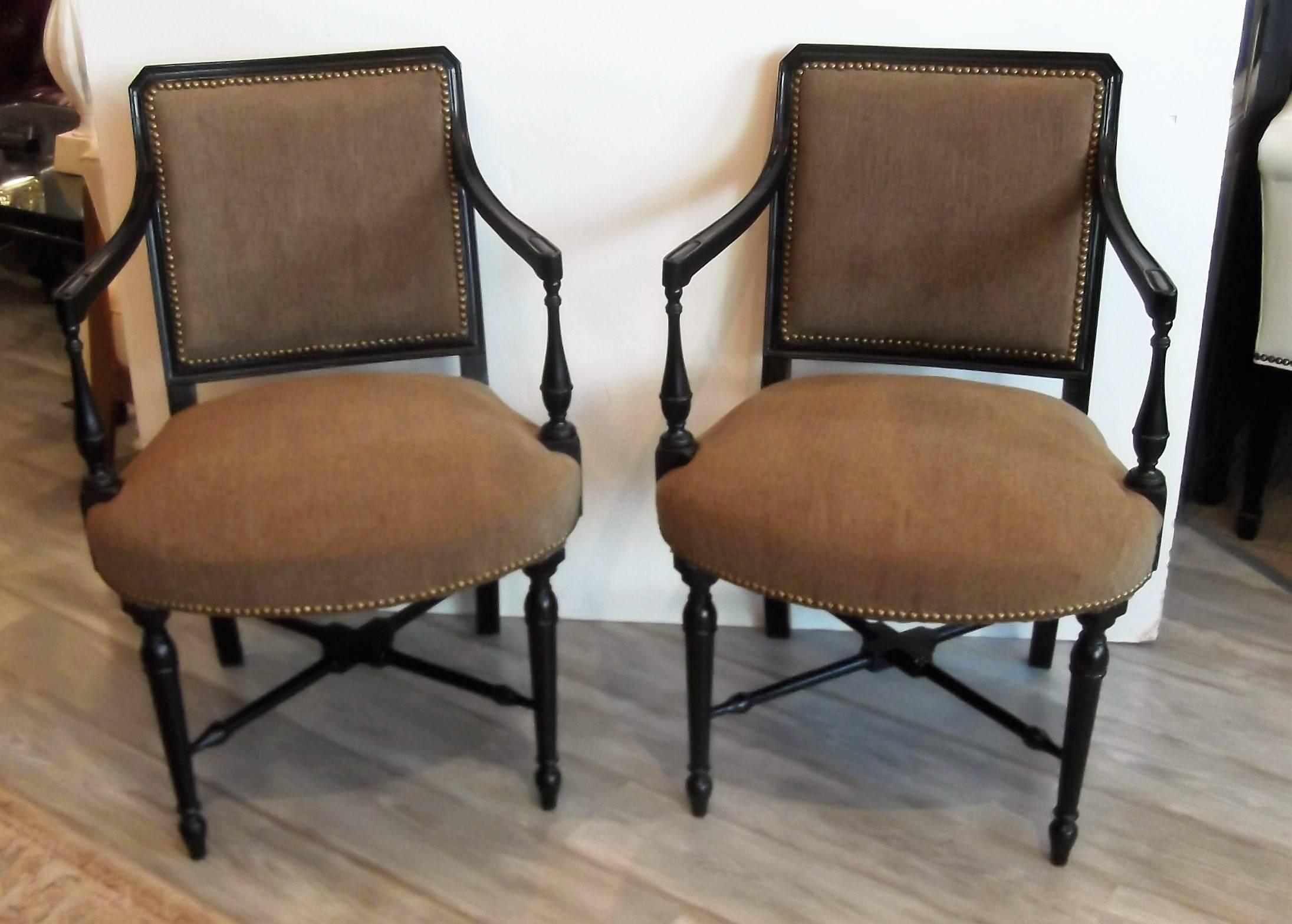 A pair of smaller ebonized armchairs with neutral olive-tan chenille upholstery. The sturdy wood frame with ebony finish is trimmed with antiqued brass nailhead trim. Nicely turned legs and X-stretcher base and slightly sloping arms.