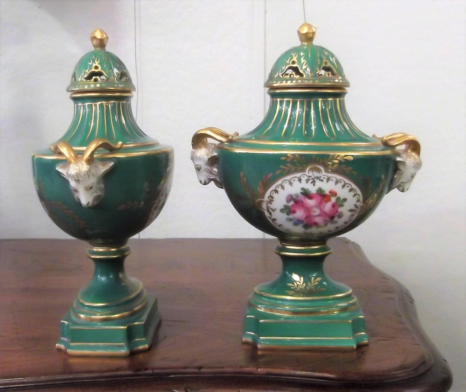 A diminutive pair of hand-painted porcelain ram's head urns with hand gilded decoration. The lids are hand pierced with small gilt finials. The center cartouches are painted Dresden flowers with a white background. The blue under glaze mark is from