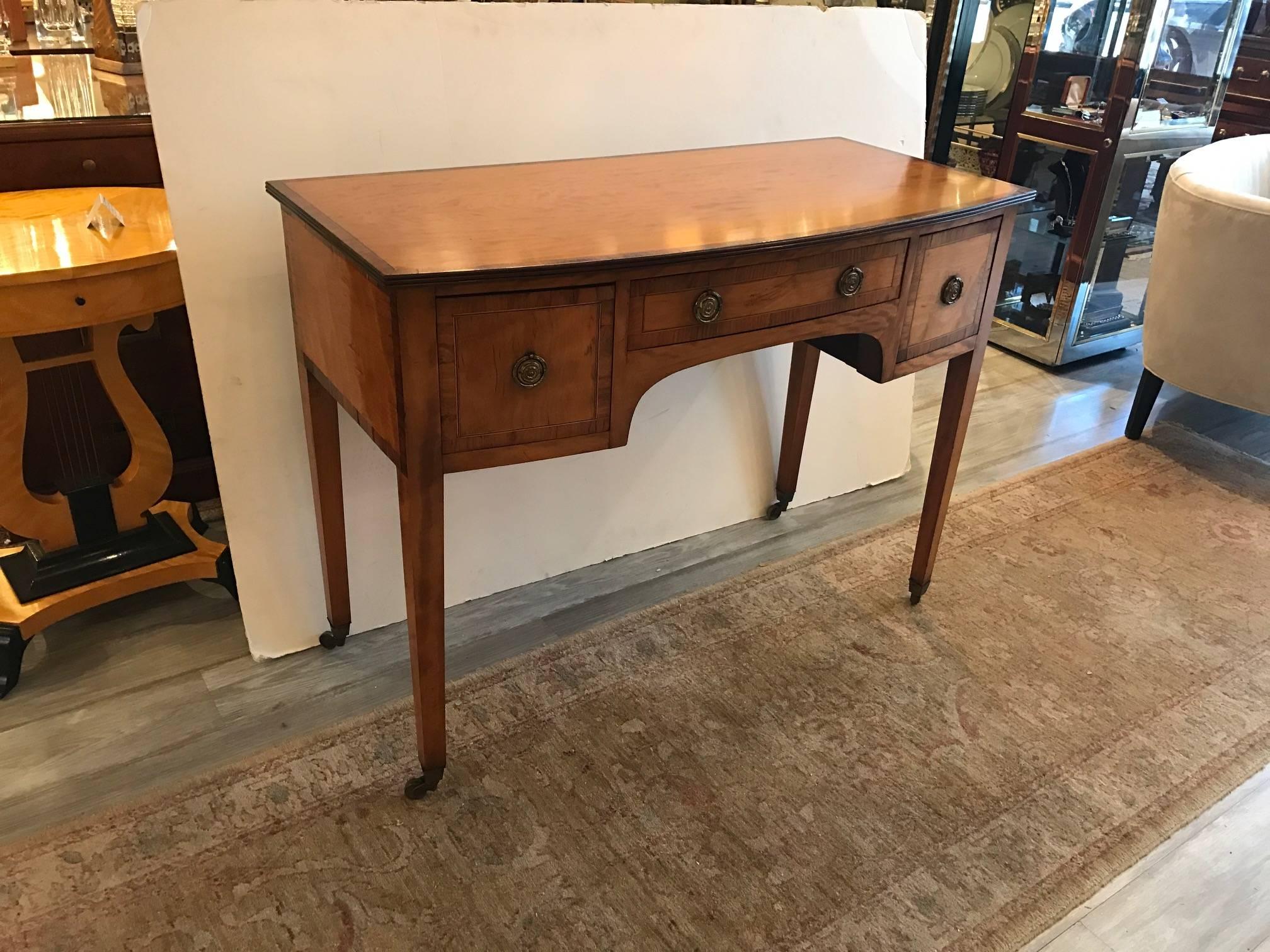 A diminutive George III satinwood bow front sideboard, the crossbanded top in rosewood with reeded edge over three similarly crossbanded drawers. all resting on square tapered legs ending in brass capped castors.