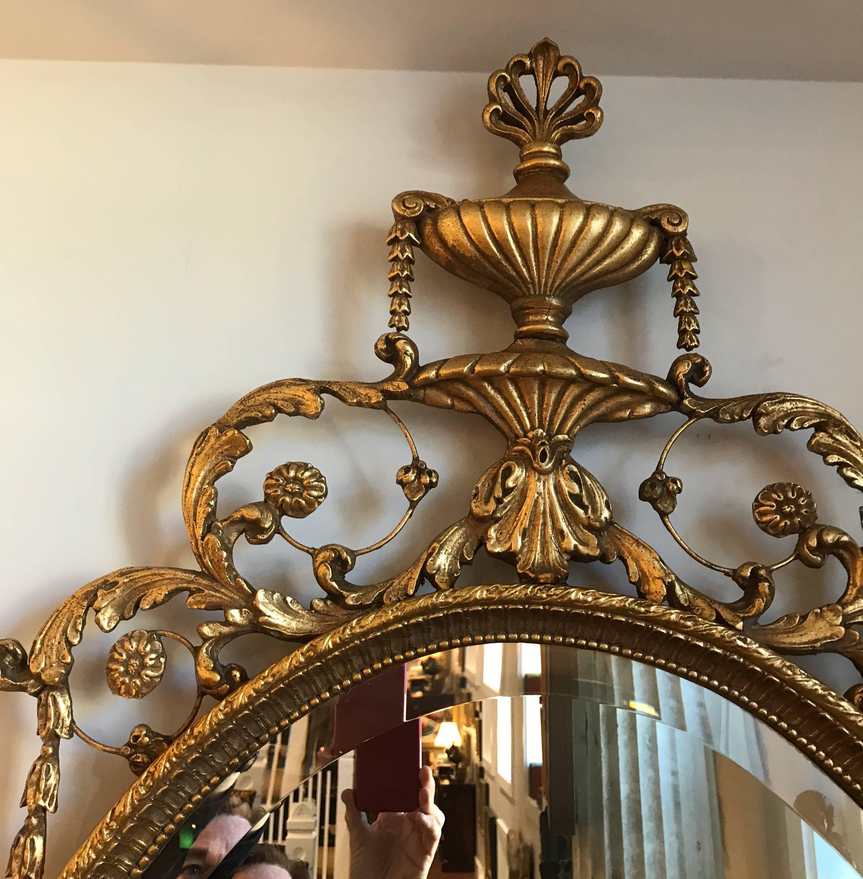 Timeless and elegant Adam style gilt wood and gesso mirror. The urn finial top with rosettes and scrolls and winged figures on each side. The bottom with swags and lower central finial. The mirror is beveled and has no silver loss and just the oval
