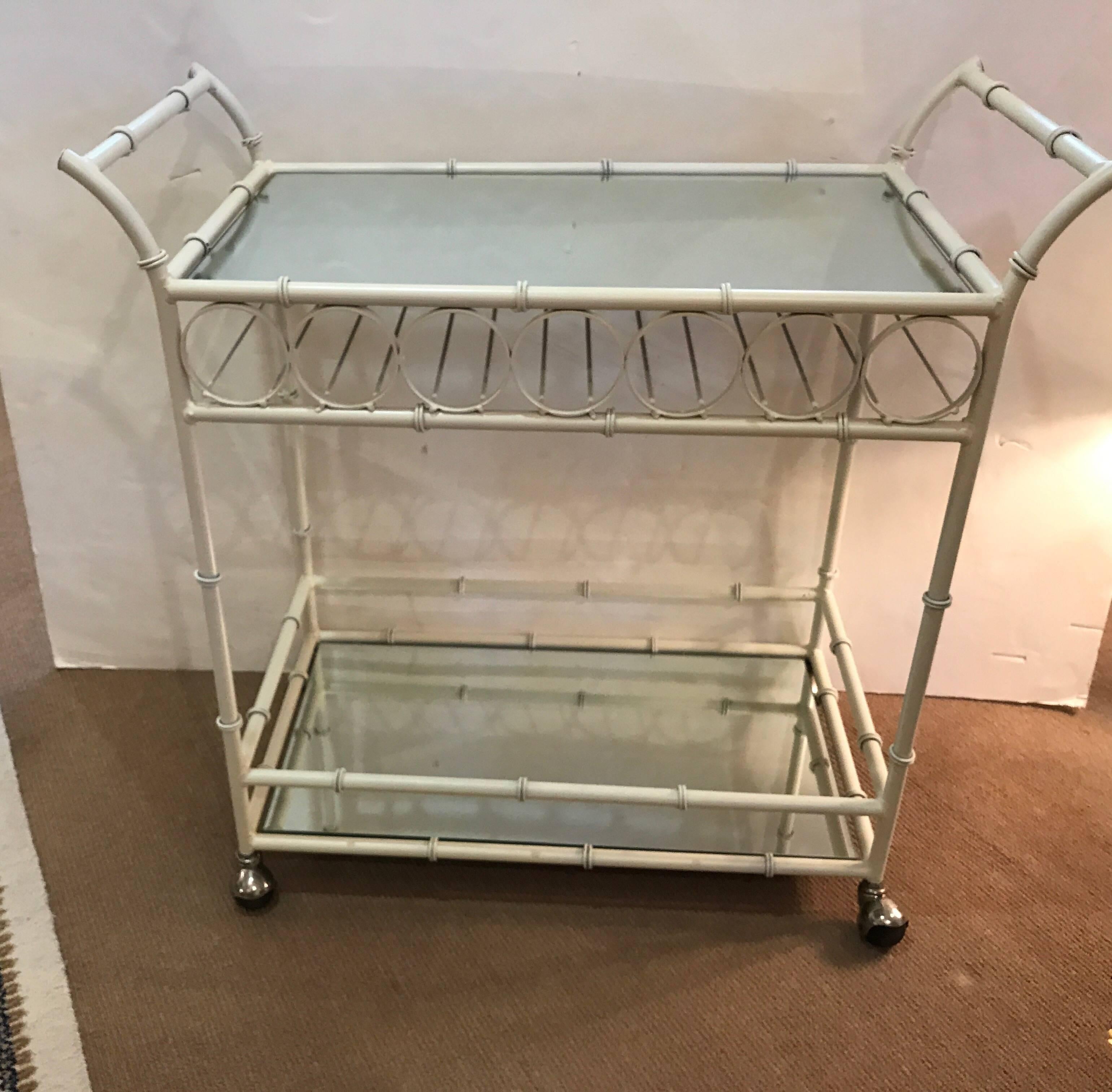 An off-white enameled metal bar cart with mirrored shelves. The flared are handles with faux bamboo motif frame. The upper shelf with storage for bottles underneath. The cart has four castors.