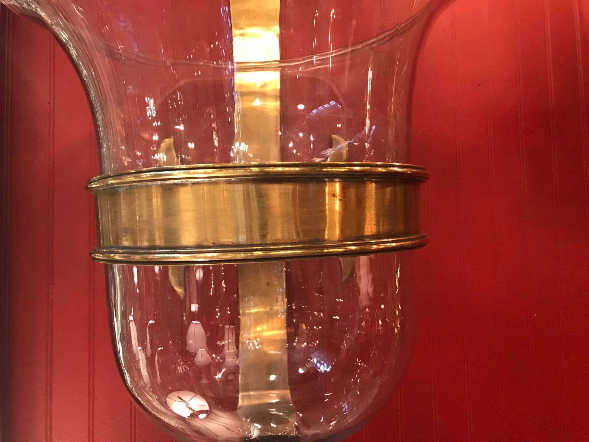 A very large pair of candle sconces with solid antique brass holders. The handblown glass hurricanes fit into broad brass rings and fit into the finialed bases. The back plates with a fleur-de-lis styled ends.