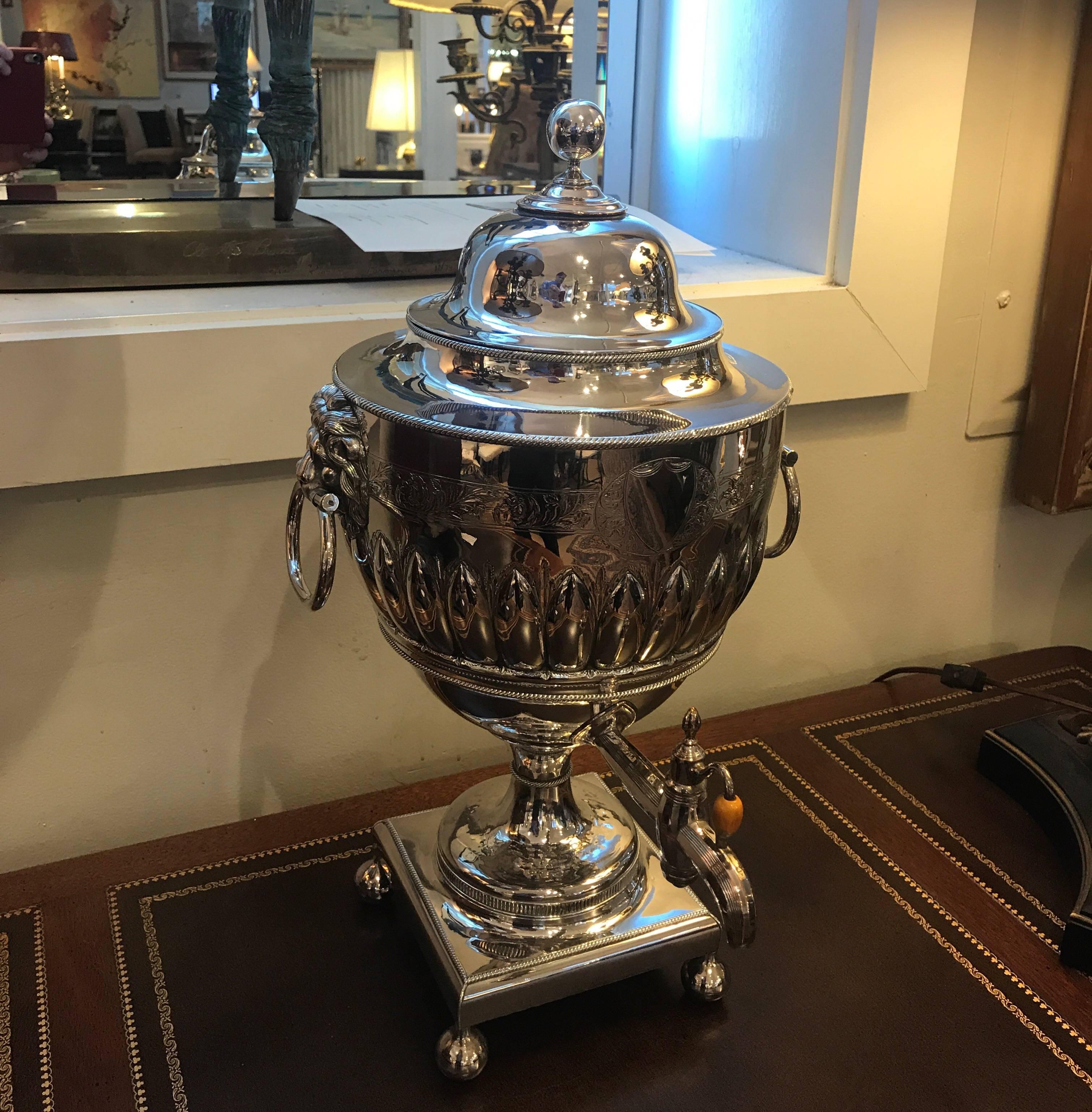 A large English silver plate tea urn with lion handles. The classic urn shape with front spigot resting on square plinth base with ball feet.