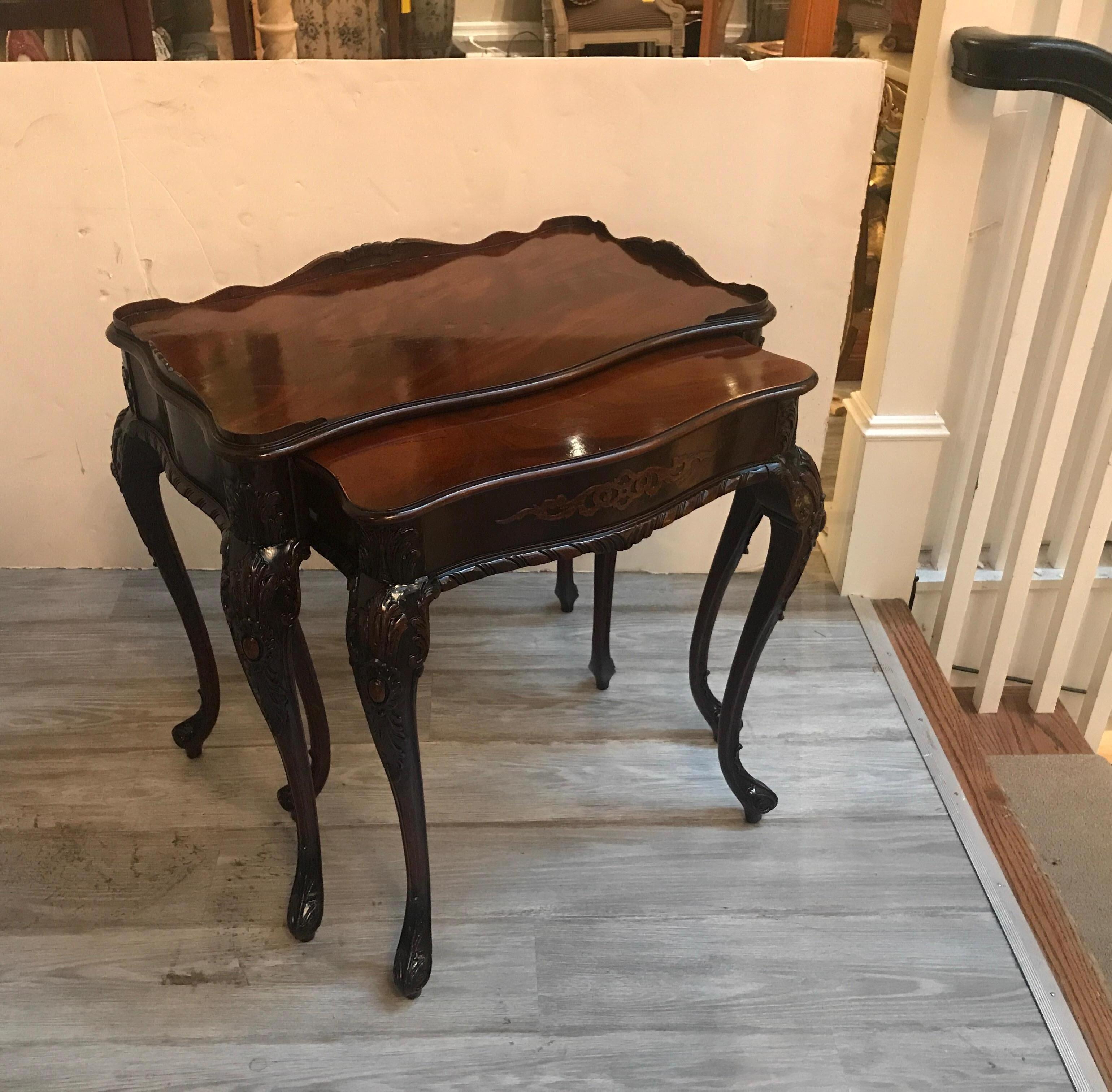 A pair of elegant hand carved mahogany nest of tables. The flame mahogany tops with gallery edge on the larger table with a smaller table that fits underneath. Beautiful carved apron and legs. These tables with recent French polish but retains some