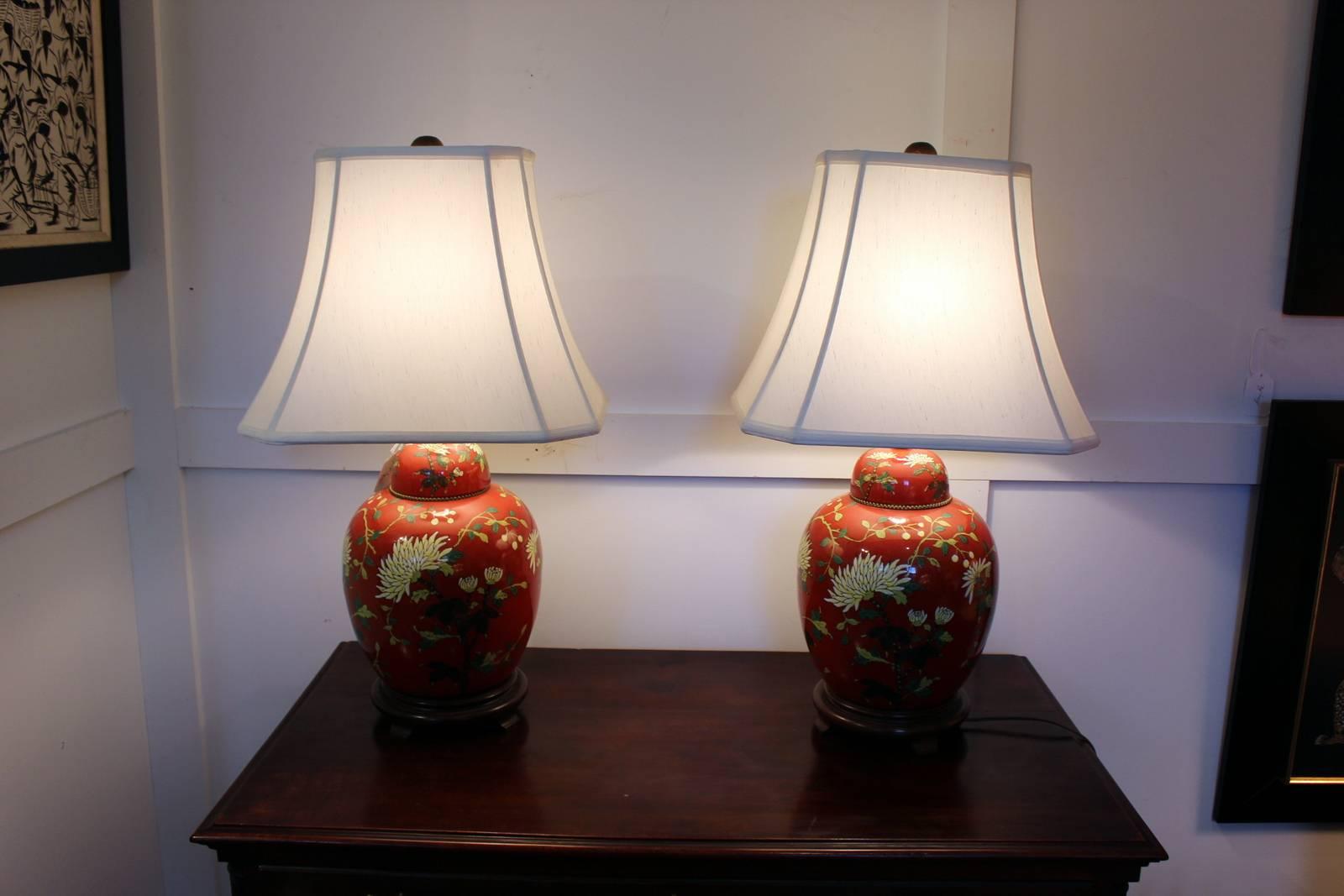 The lamps with iron red background with raised enameled floral decoration rest on wood bases. The wood base with diminutive feet. The shades are for photographic purposes only and not included with these lamps. Light chip on the wood bases.