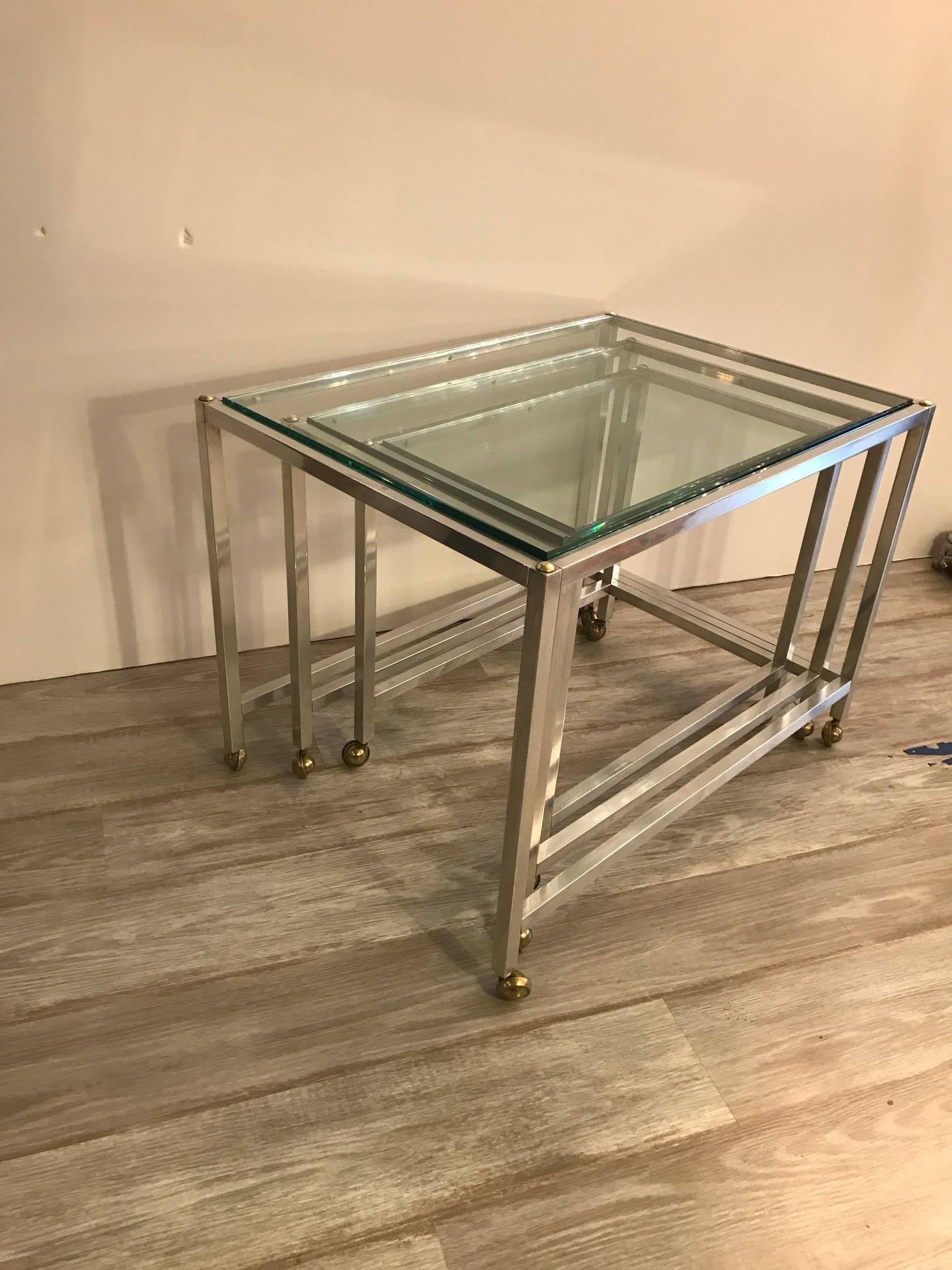 Mid-Century Modern set of glass top nesting tables. The cube form of three tables with castors, each table fits neatly into the other in sturdy polished aluminium with brass appointments. The largest table is 20 by 28, 20 inches tall, the smallest
