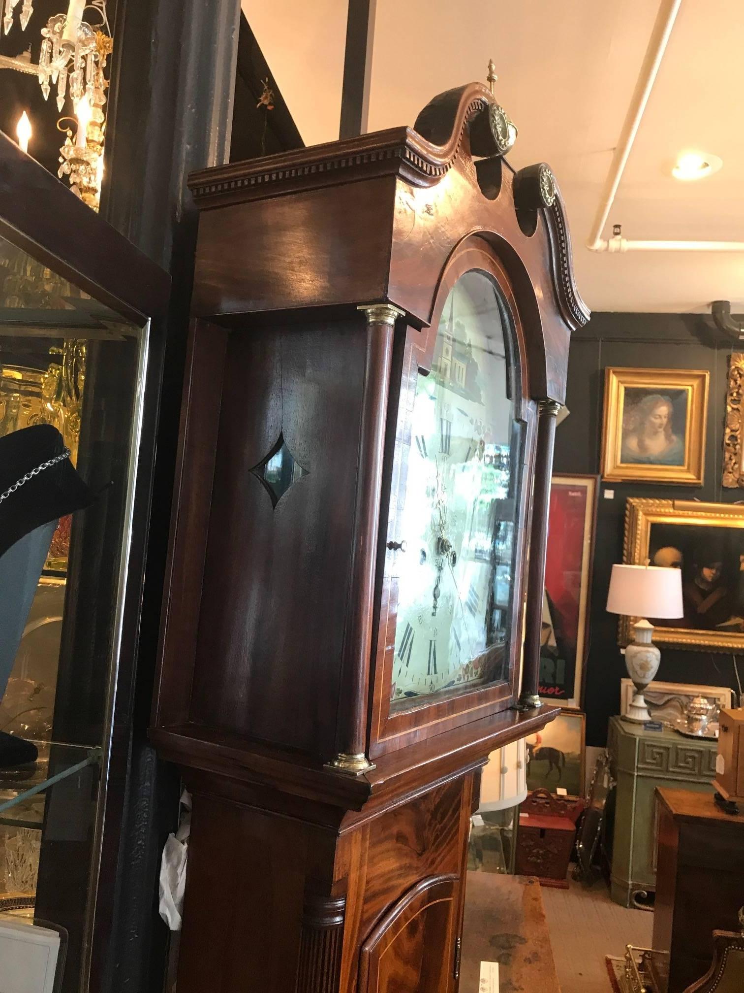 George III mahogany tall case clock, circa 1800. The bonnet with a swans neck pediment and dental molding with central finial, the sides with Doric columns with brass capitals flanking a glazed door opening to a white painted dial. The clock is