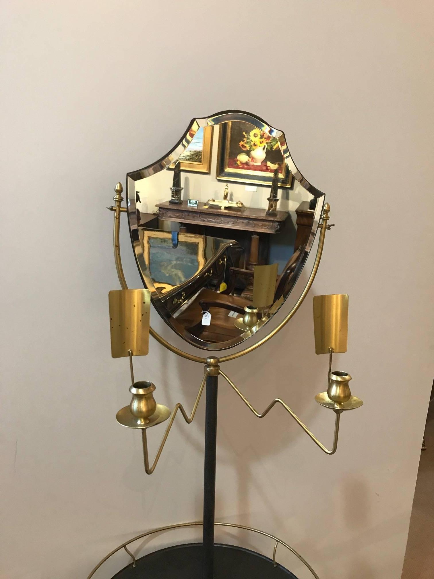 Elegant brass and galvanized metal standing shaving or dressing mirror. The Classic shield shaped mirror flanked by a candleholder with reflector. The center with a gallery edge table resting on a tripod base.
