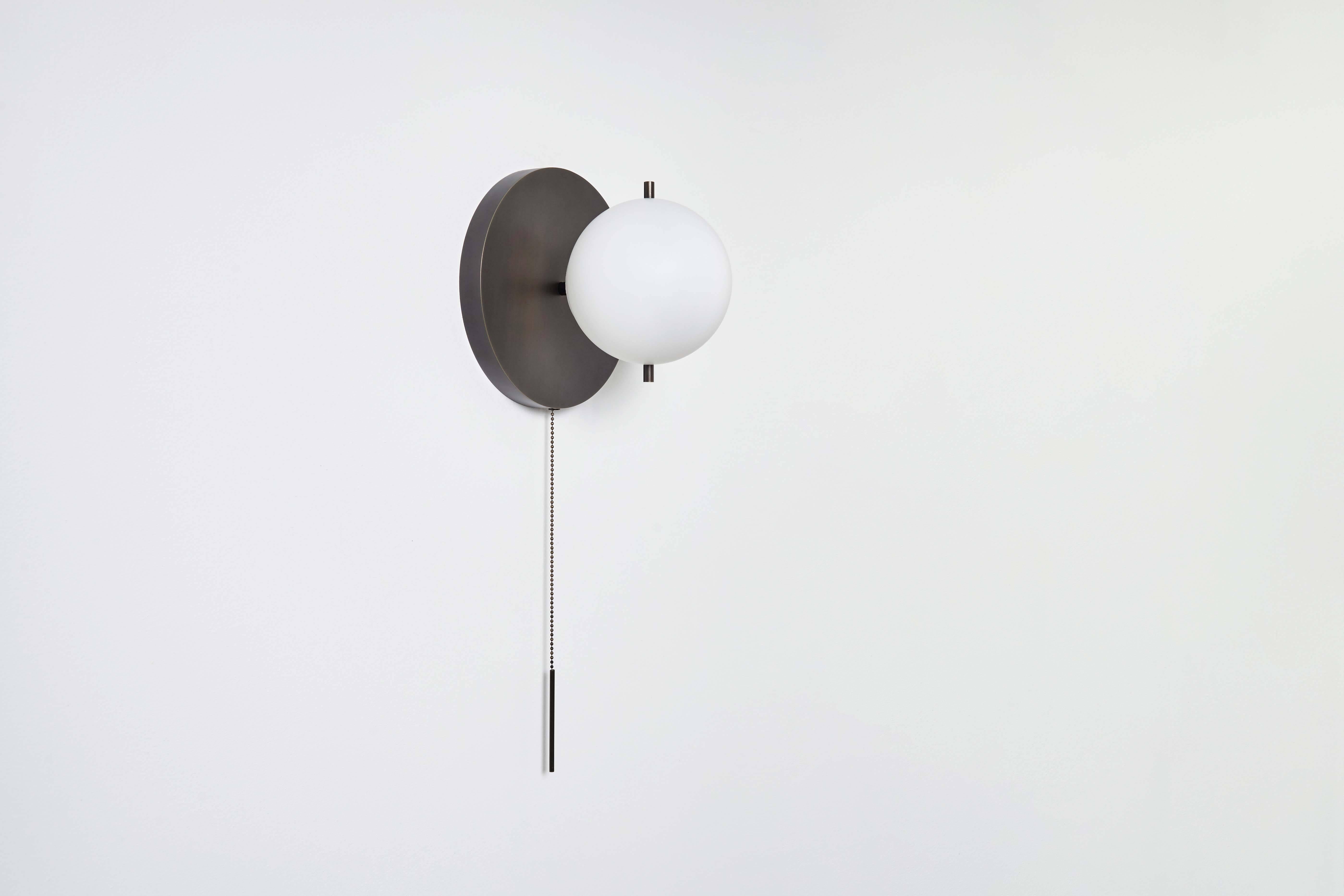 The signal sconce features a hand sanded globe. The fixture is delicately mounted between two metal pins above a luminous canopy, creating a hieroglyphic composition. A pull chain holds a slender rod, giving the piece a jewel-like presence.