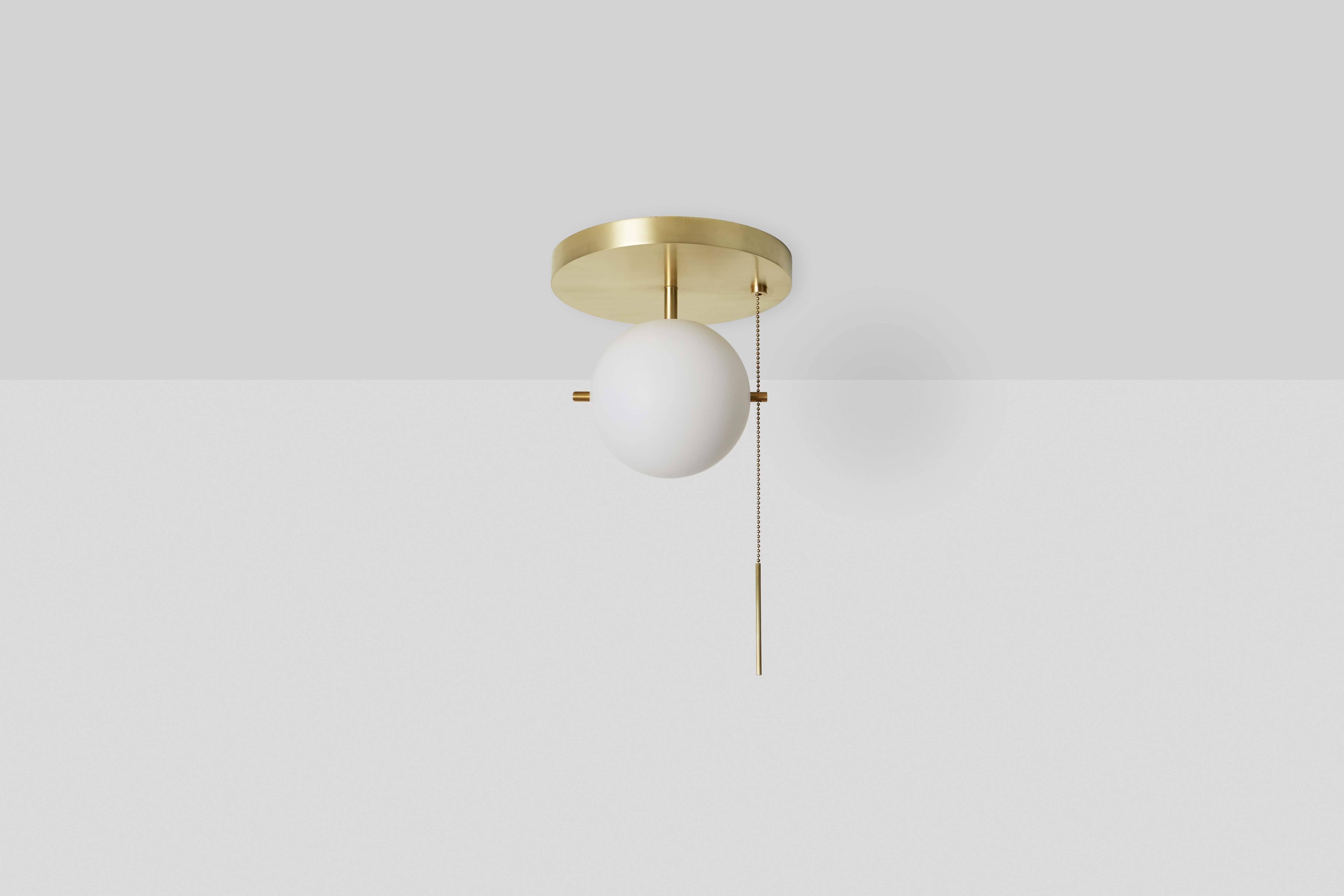 The Flush Mount is a compact beacon composed of metal, glass, and chain. A hand blown glass globe is delicately mounted between two metal pins above a luminous canopy, creating a hieroglyphic composition. The pull chain holds a slender rod, giving