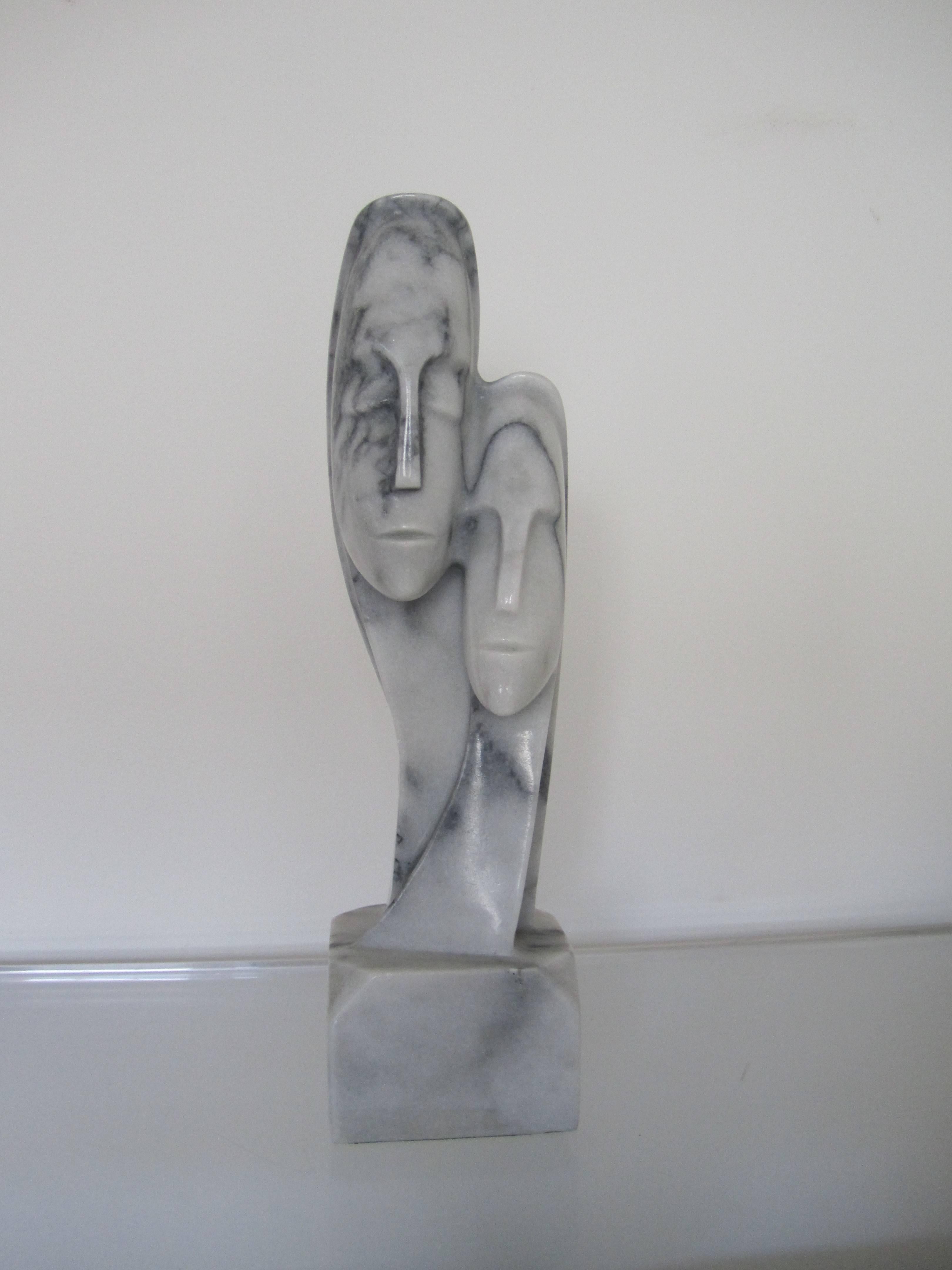 Importante escultura figurativa tallada en mármol, de estilo posmoderno o moderno, con la cabeza y el busto de dos personas abrazadas, de mediados a finales del siglo XX. El mármol es gris con vetas oscuras. La pieza escultórica está tallada en una