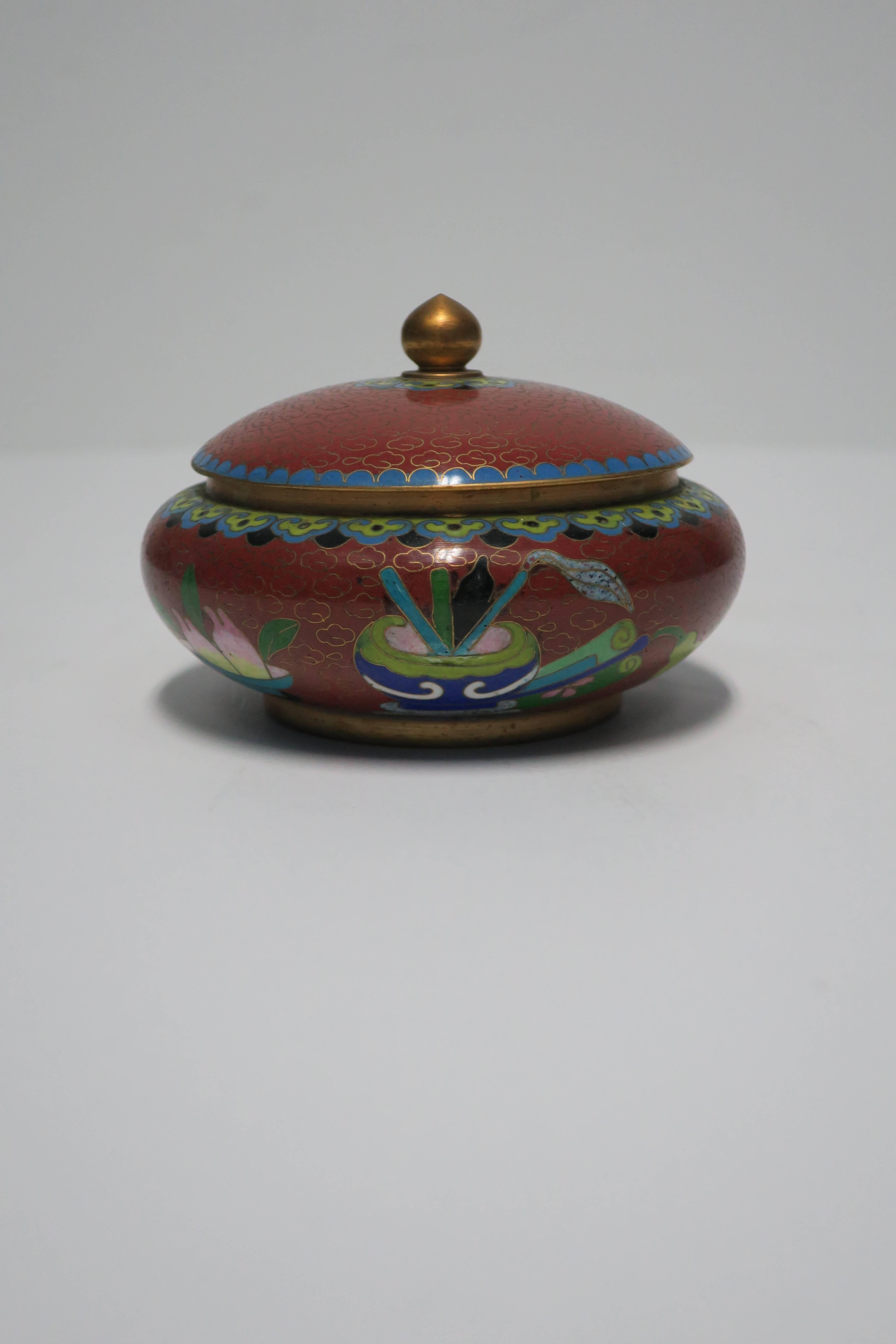 A beautiful vintage Chinese cloisonné round box with brass finial top, with an exterior depicting a humming bird and blossoms. Predominant color is a dark red, burgundy, or ox blood hue, with blue, black, green, pink, and cream accent enamel
