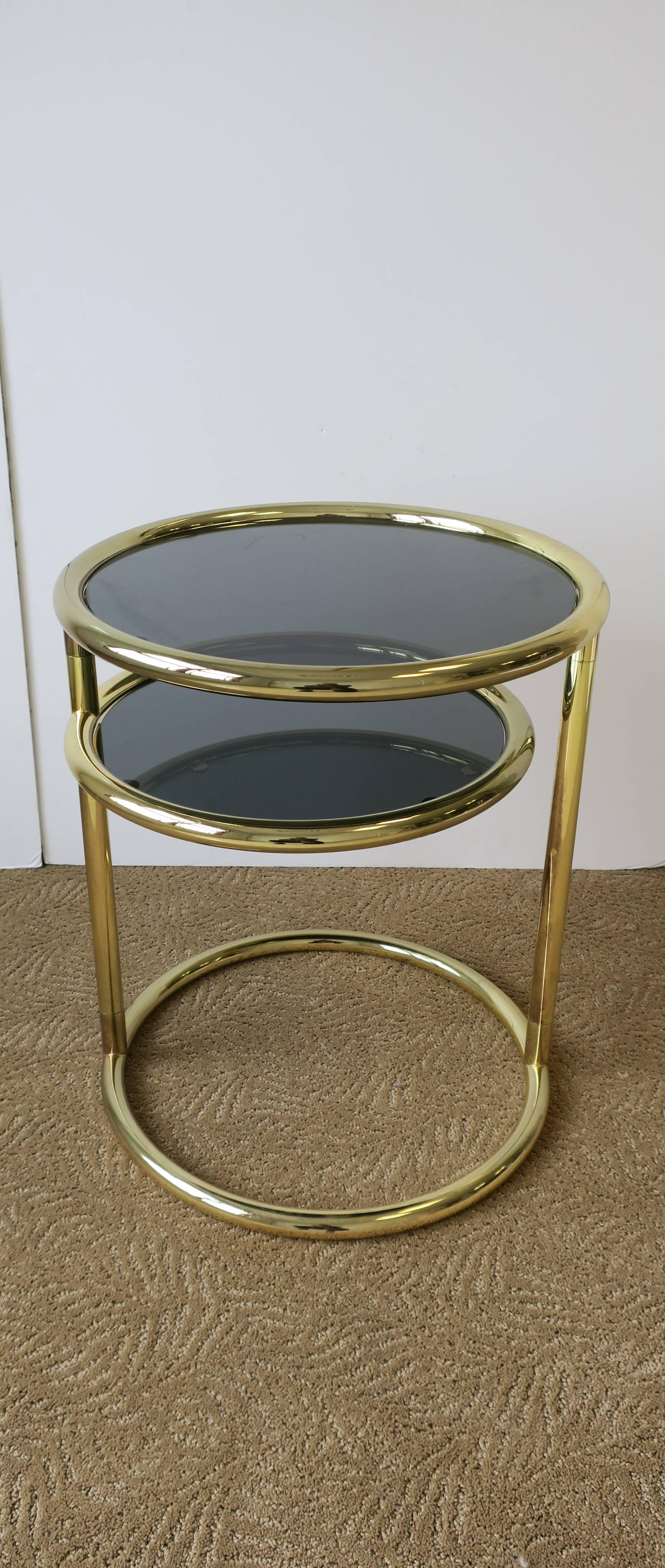 A '70s Modern tubular brass and smoked glass round side table with swivel shelf. Side table has a lower shelf that swivels out, as demonstrated in images. Table is in the style of designer Milo Baughman, circa 1970s. 

Measurements include: 21 in. H