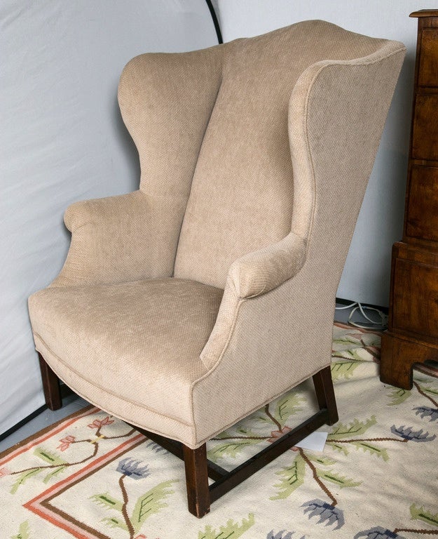 This wingback chair with a mahogany frame sits on raised square legs joined by stretchers.  The chair has been reupholstered in beige figured velvet.