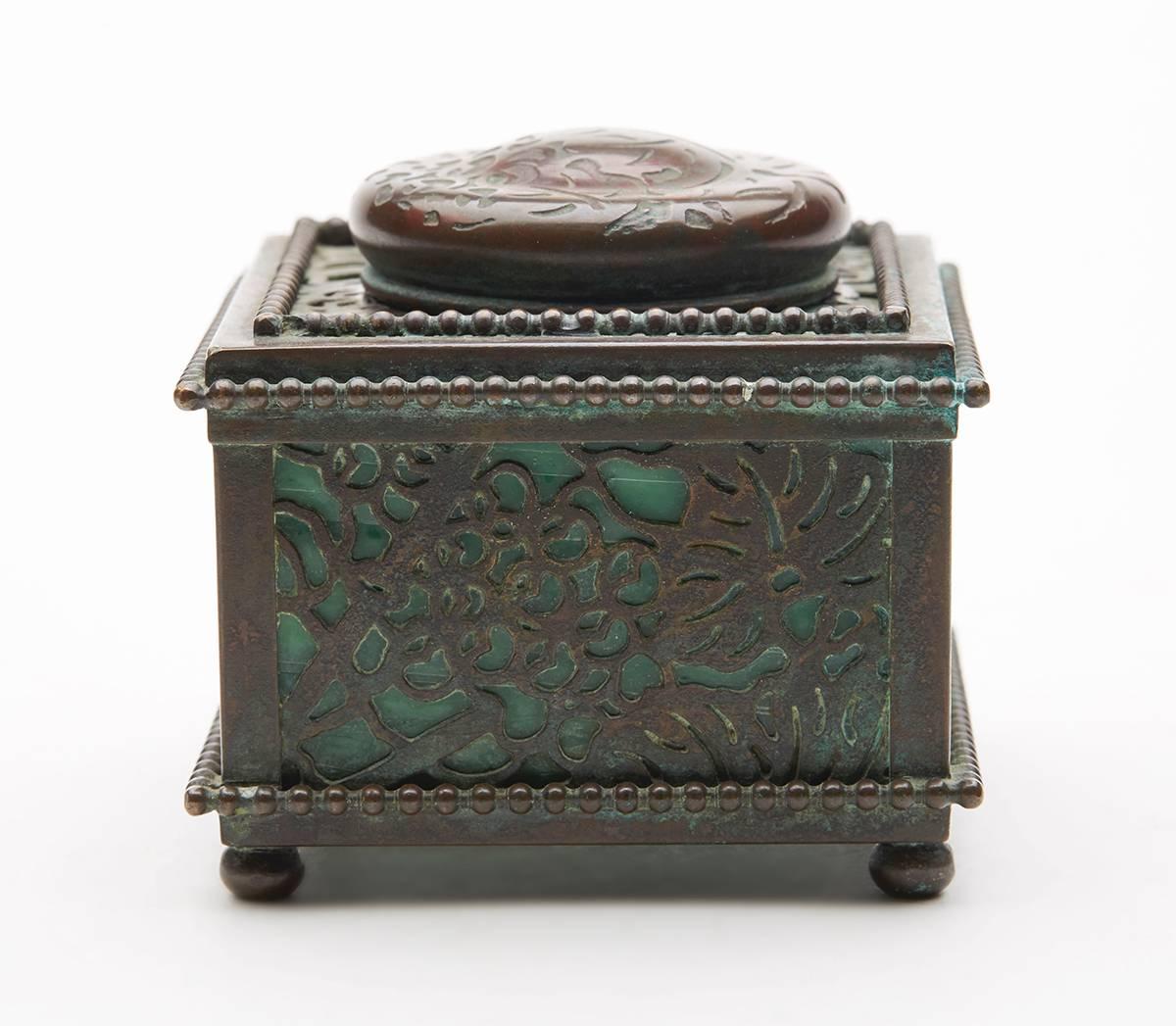 A fine antique Tiffany Studios bronze mounted marbled green glass lidded inkwell in the grape pattern design. The square shaped inkwell stands mounted on four ball feet with open work panels with grape vine designs over green marbled glass panes.