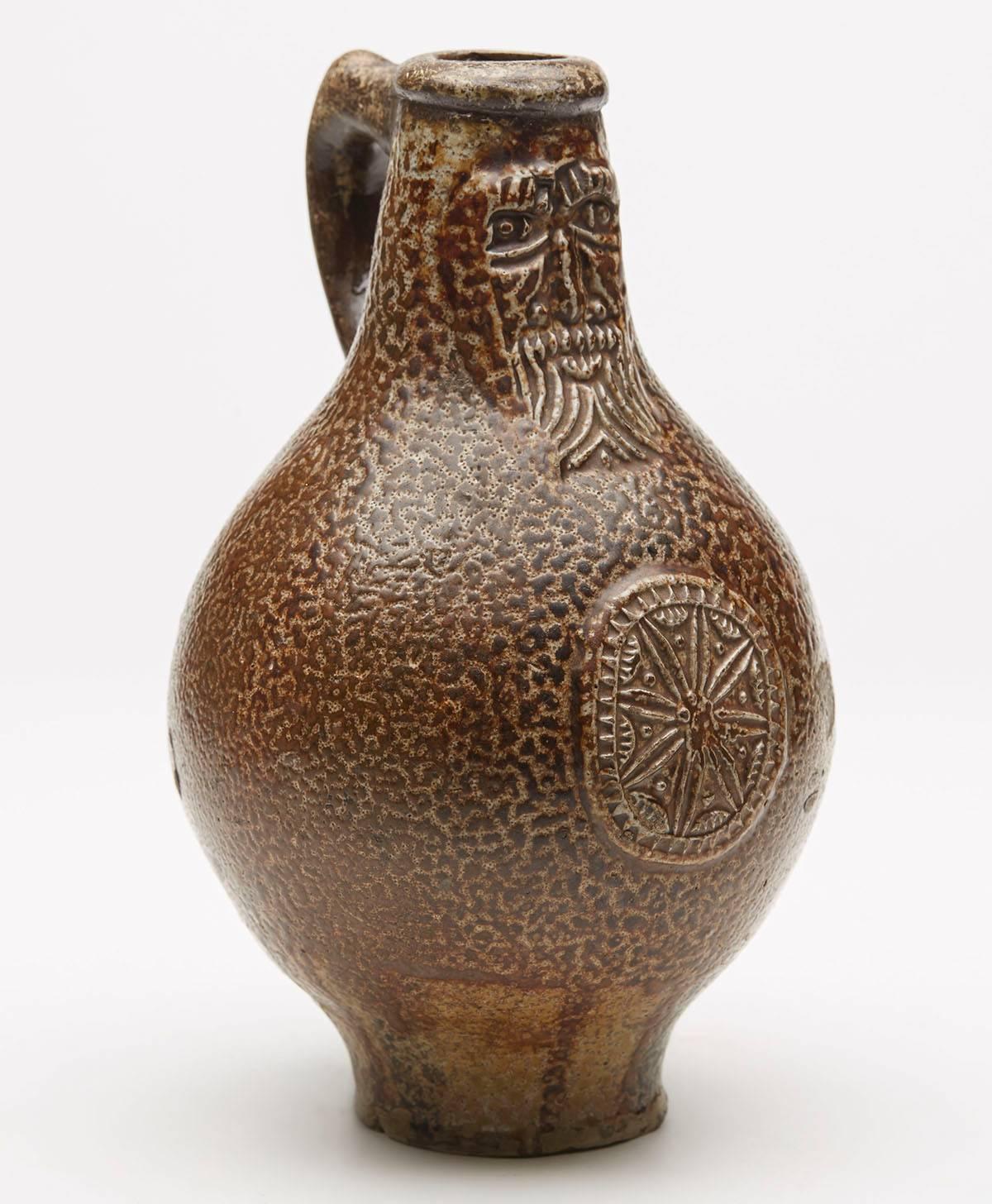 An interesting antique German Rheinish Bellarmine bottle shaped stoneware jug applied with a bearded mask along with a floral crest to the body. The jug has a brown salt glazed speckled finish and is not marked.
   