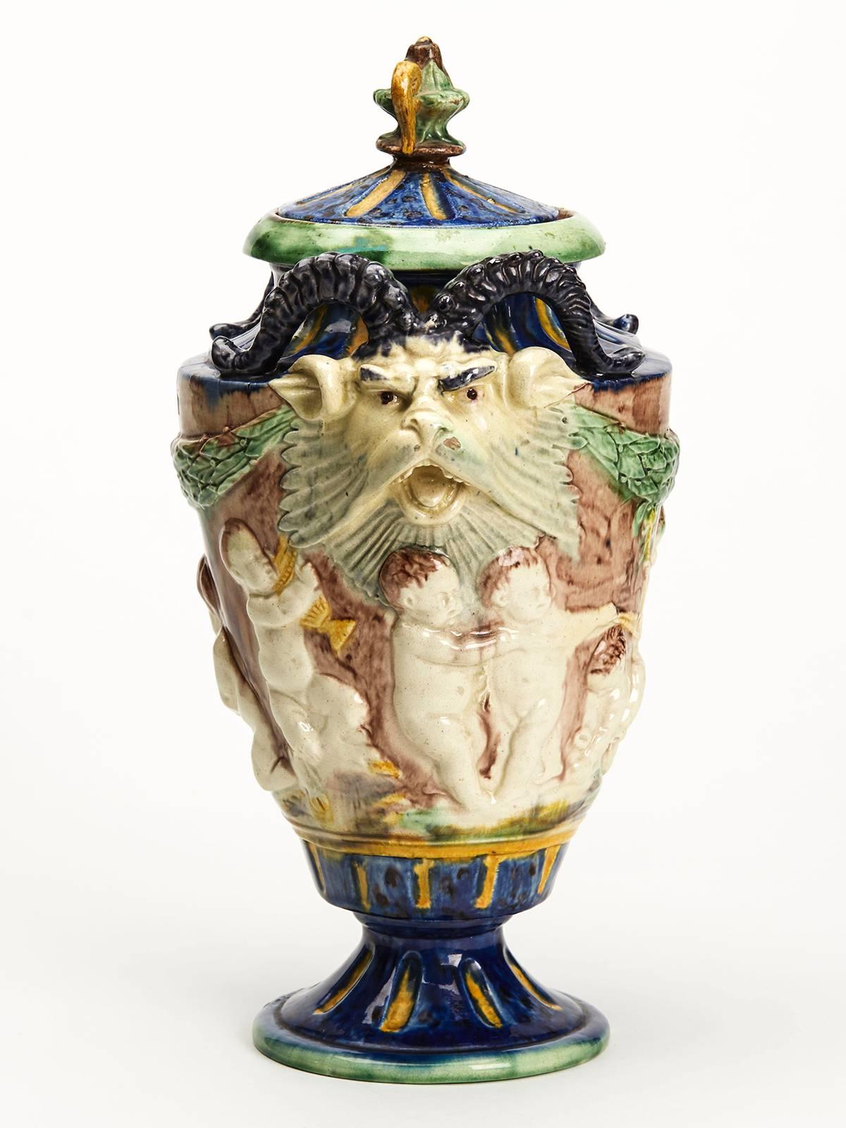 An antique Majolica lidded vase moulded with a continuous relief moulded scene of putti in various activity with grotesque horned lion heads applied either side of the rim. The earthenware vase stands on a rounded pedestal foot with a rounded raised