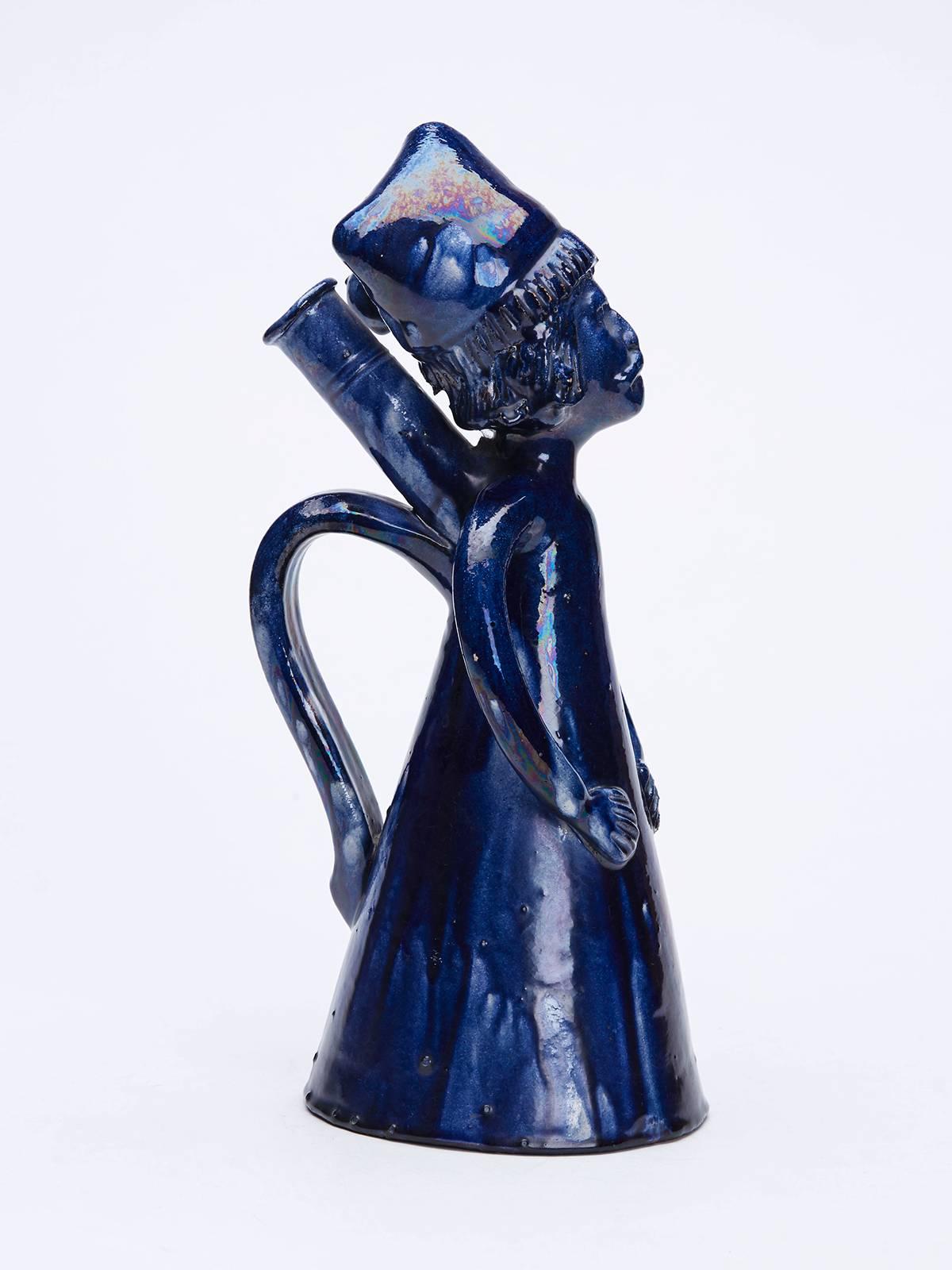 Un ancien pichet ou flacon d'art populaire en poterie de terre rouge émaillée bleue, représentant un homme portant une chemise de nuit et un bonnet de nuit, avec un bec verseur derrière la tête de l'homme, une poignée de transport en dessous et des