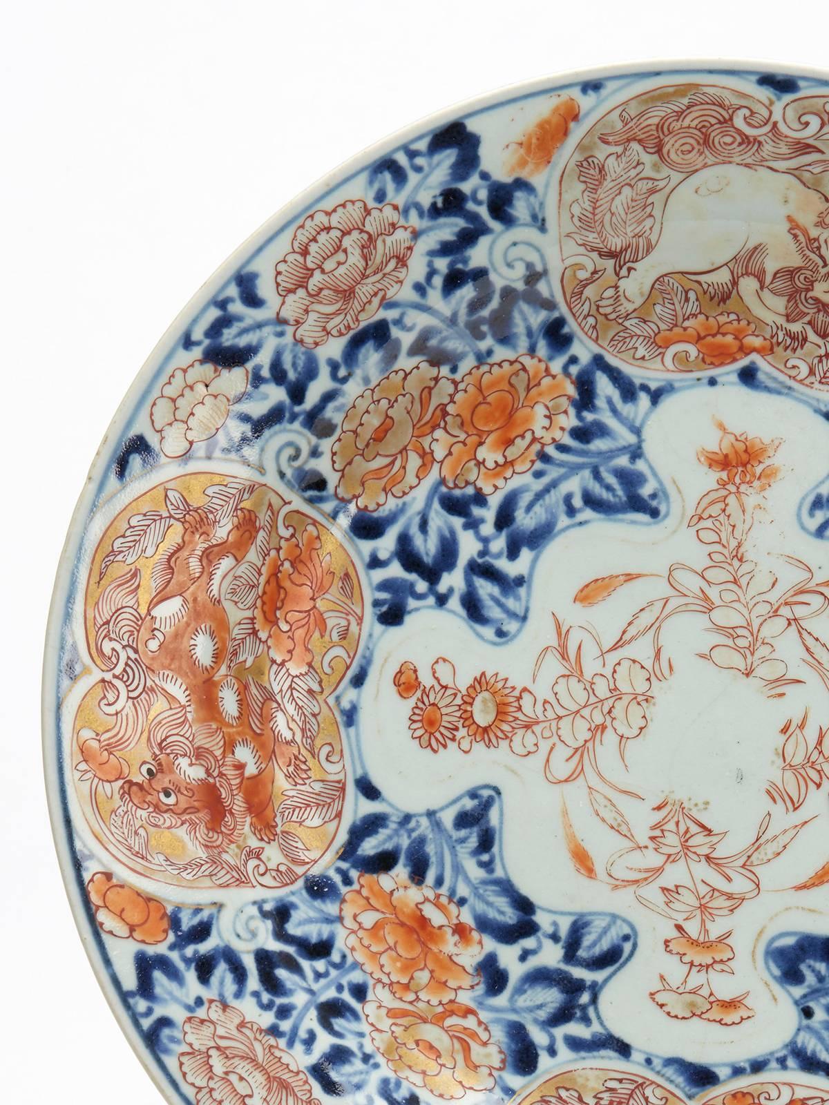 Eine japanische Porzellanschale aus der Meiji-Zeit, dekoriert im alten Imari-Stil mit Tafeln, die mythische Tiere zeigen. Die runde, flache Schale ist mit blauer Unterglasurmalerei und roter Übermalung mit Vergoldung verziert. Auf vier Feldern sind