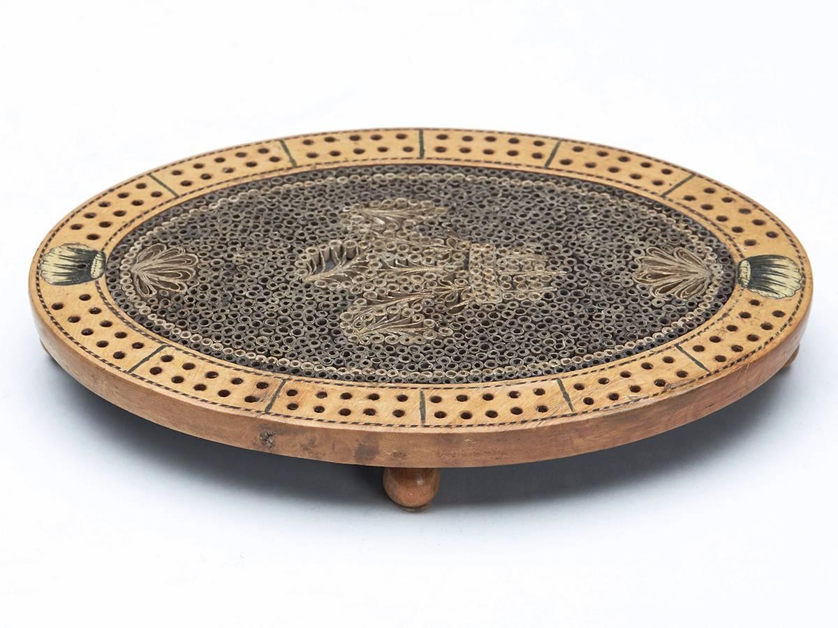 A very rare Georgian oval wooden cribbage board with inlaid quillwork design incorporating Prince of Wales feather design. The board stands raised on four ball feet with circular lidded peg well to the base and with drilled edge with painted designs