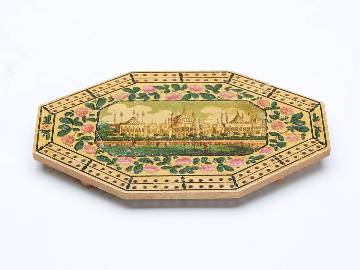 A very rare Georgian octagonal wooden cribbage board with a central framed scene of Brighton Pavillion within a floral border with peg holes arranged around the outer edge within simple painted line borders. The cribbage board stands raised on four