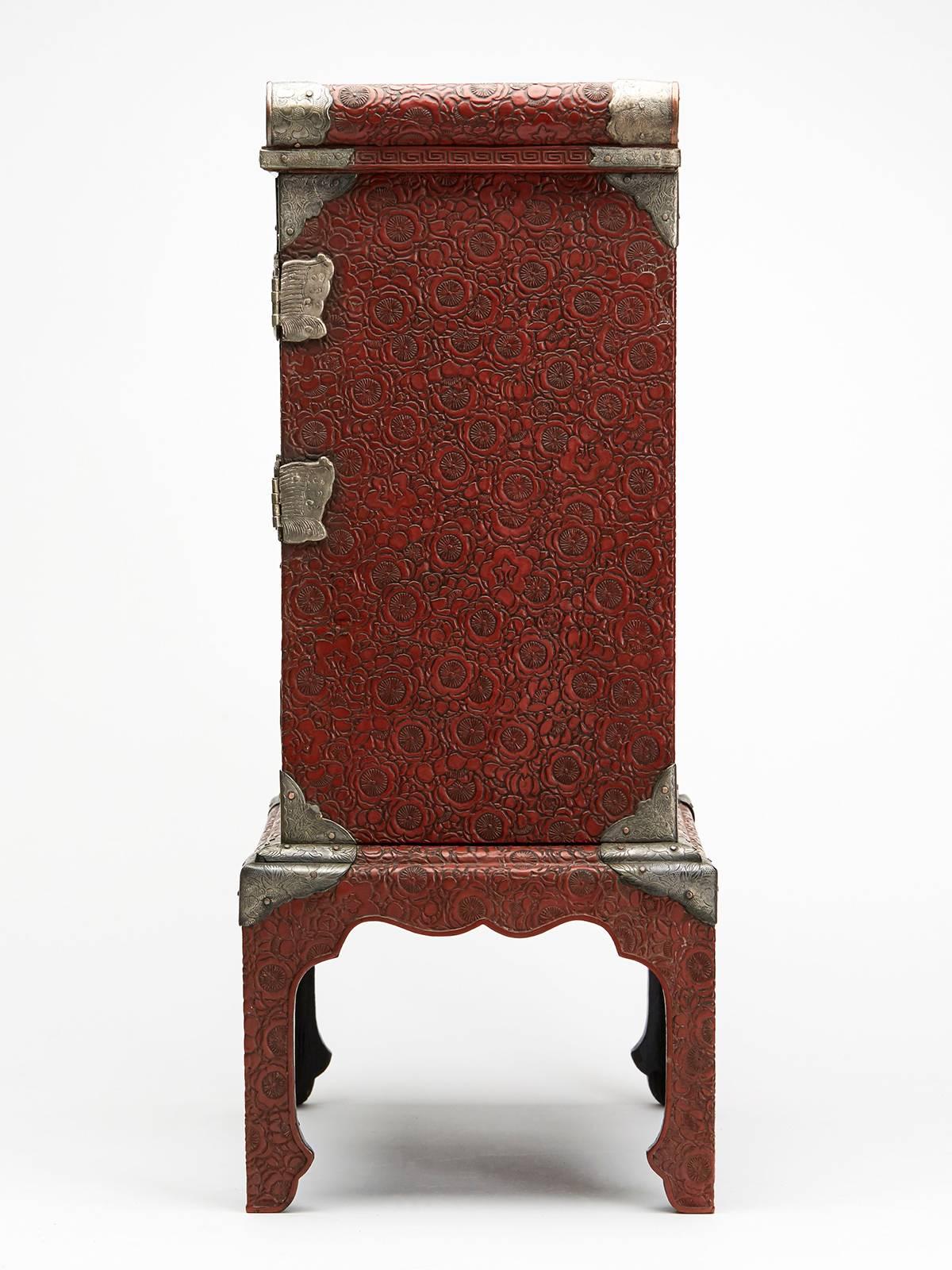An exceptional antique Japanese red cinnabar lacquer table cabinet on stand inset with finely painted Satsuma panels, possibly Kinkozan, with engraved metal mounts with locking doors and drawer. This elegantly shaped cabinet has a pagoda shaped top