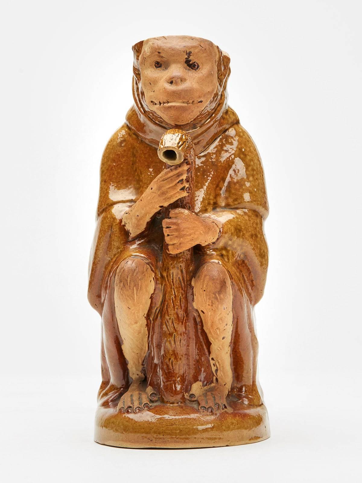 An unusual antique salt glazed pottery seated monkey teapot or jug portraying a monkey wearing a monks gown seated and resting on a walking stick which forms the spout. The monkey's tail forms a loop handle and is decorated in a brown salt glazed