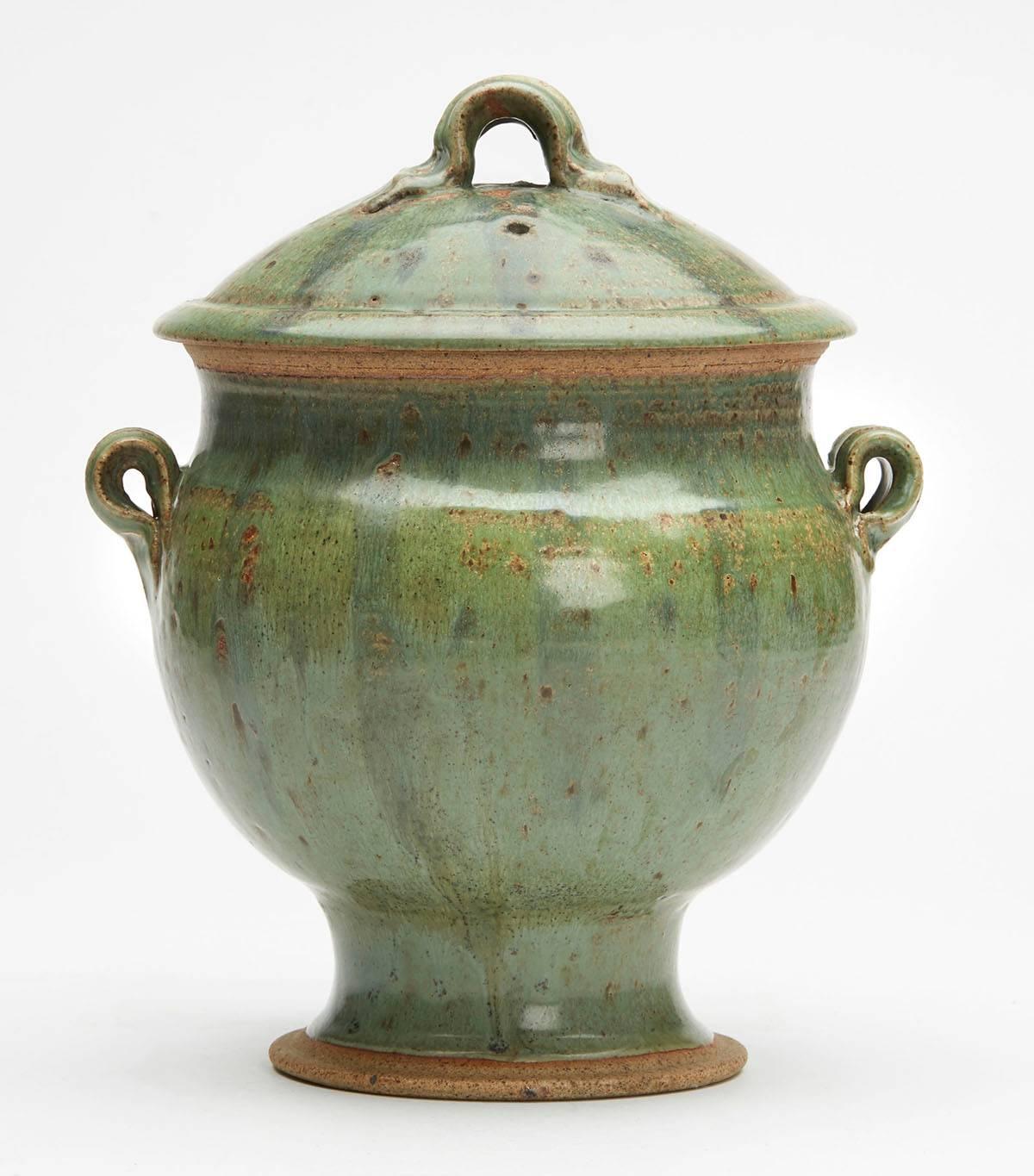 A vintage Studio Pottery twin handled lidded stoneware urn decorated in mottled green glazes. The urn stands on a rounded pedestal foot with an unglazed rim with a rounded bulbous body with small looped ridged handles and with a hat shaped cover